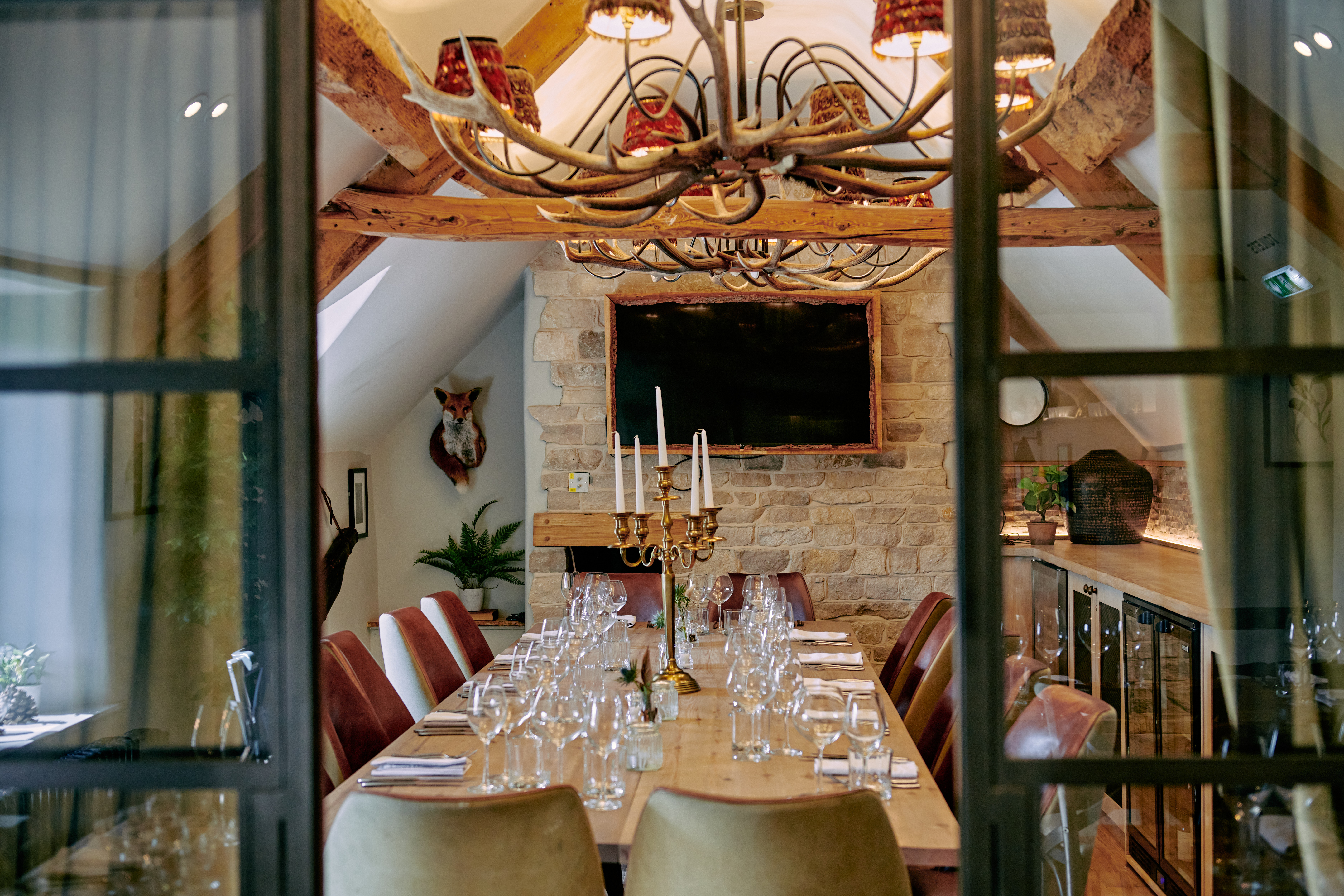 Elegant dining room with a long wooden table, set for a meal. Antler chandelier and stone wall with TV. Rustic decor. - Wild Thyme & Honey Hotel in the Cotswolds