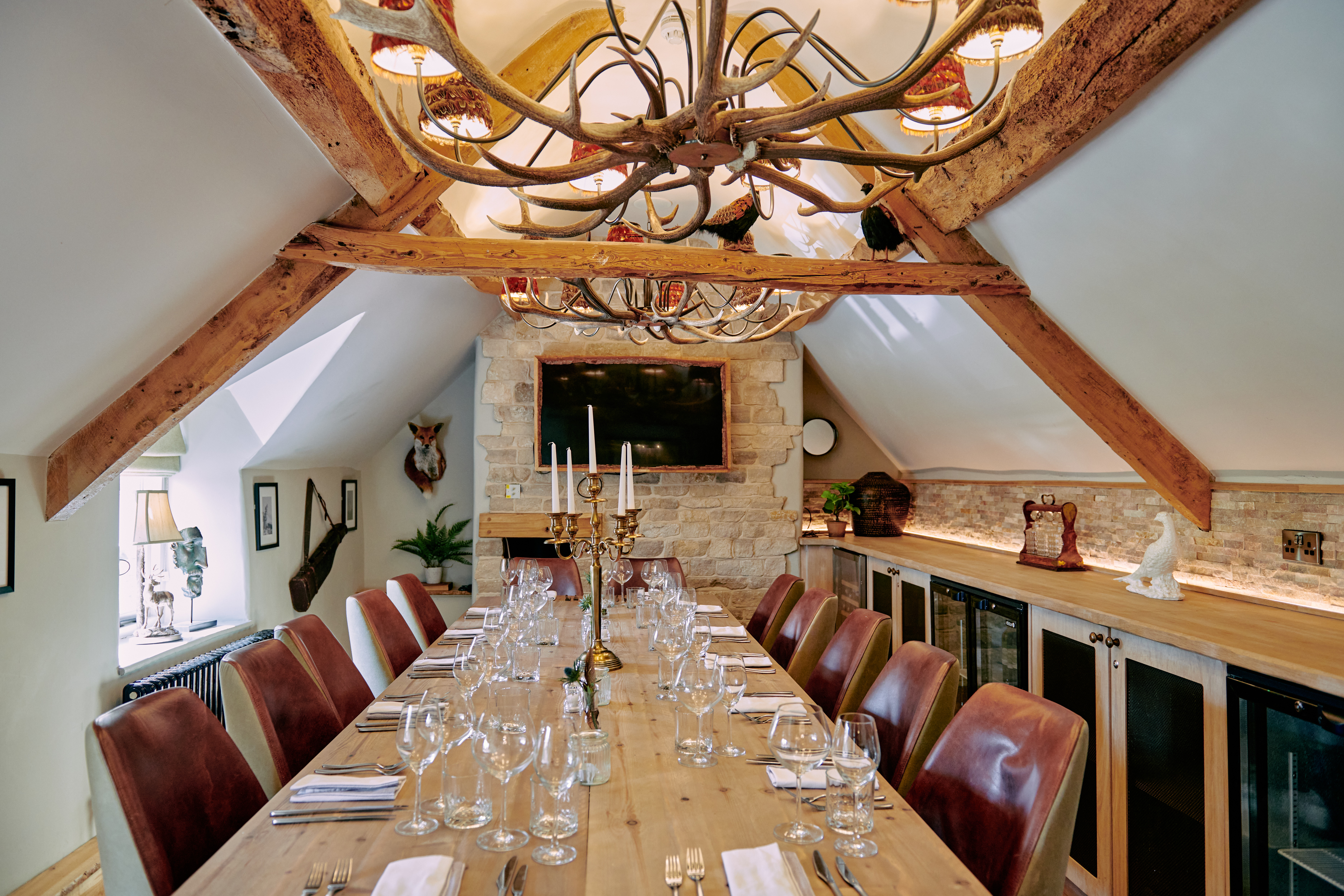 Rustic dining room with a long wooden table, leather chairs, antler chandelier, sloped ceilings, and stone accent wall. - Wild Thyme & Honey Hotel in the Cotswolds