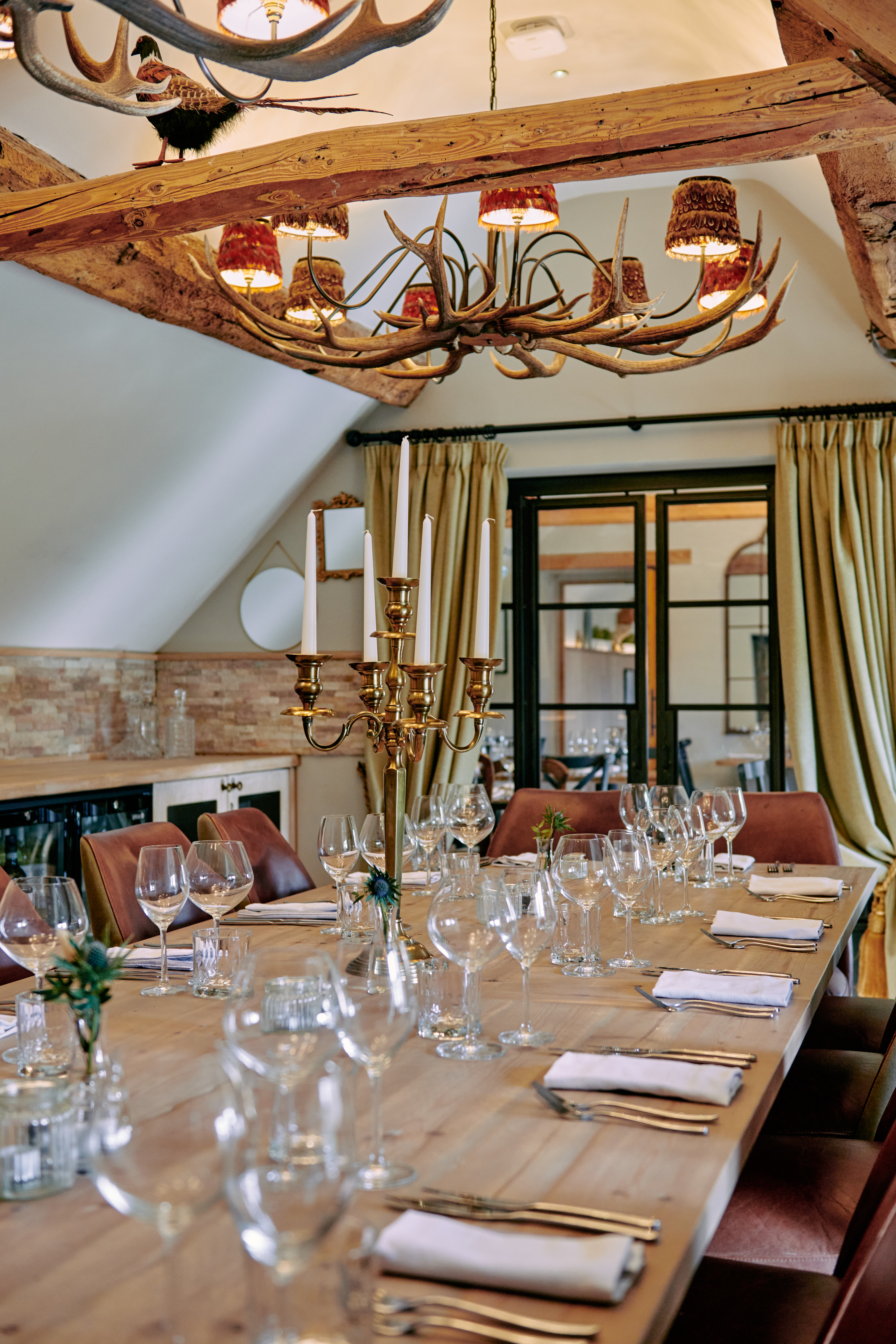 Elegant dining room with a long wooden table set with glassware, napkins, and candles, under rustic chandeliers and beams. - Wild Thyme & Honey Hotel in the Cotswolds