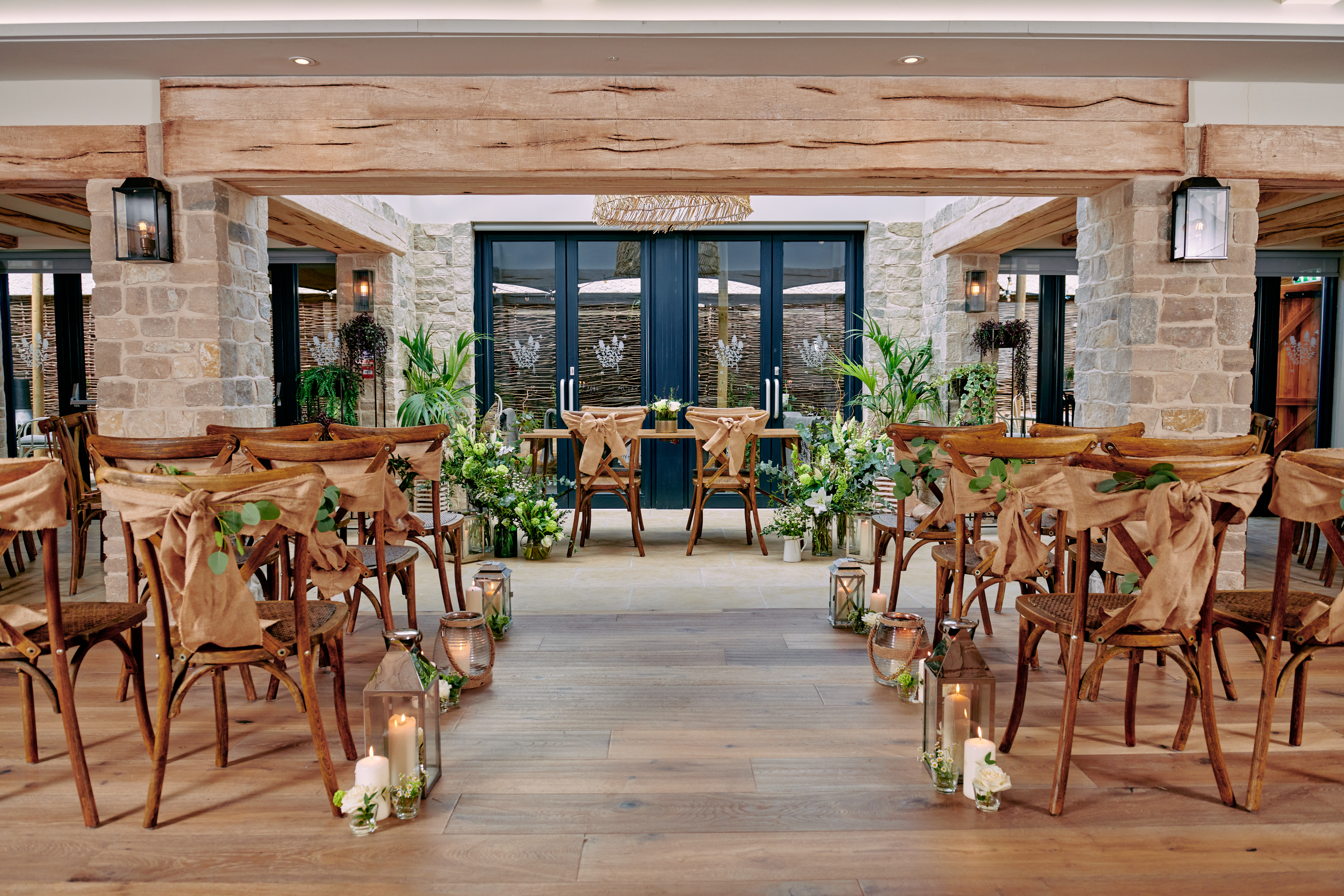 Rustic indoor wedding setup with wooden chairs, greenery, lanterns, and candles, arranged along an aisle leading to a floral arch. - Wild Thyme & Honey Hotel in the Cotswolds