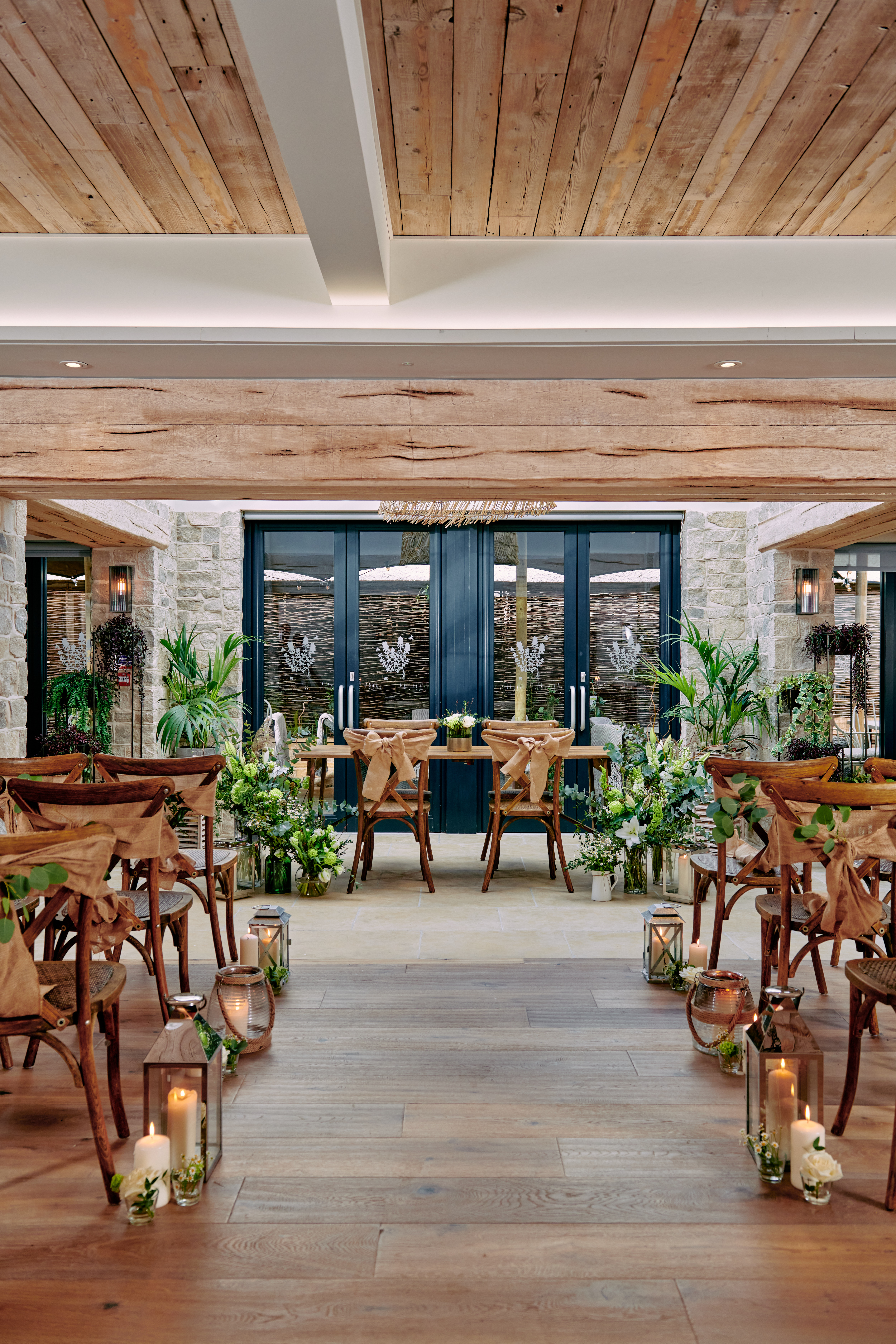 Elegant indoor wedding venue with wooden chairs, floral decorations, and candles lining the aisle. Tables at the end, large windows behind. - Wild Thyme & Honey Hotel in the Cotswolds