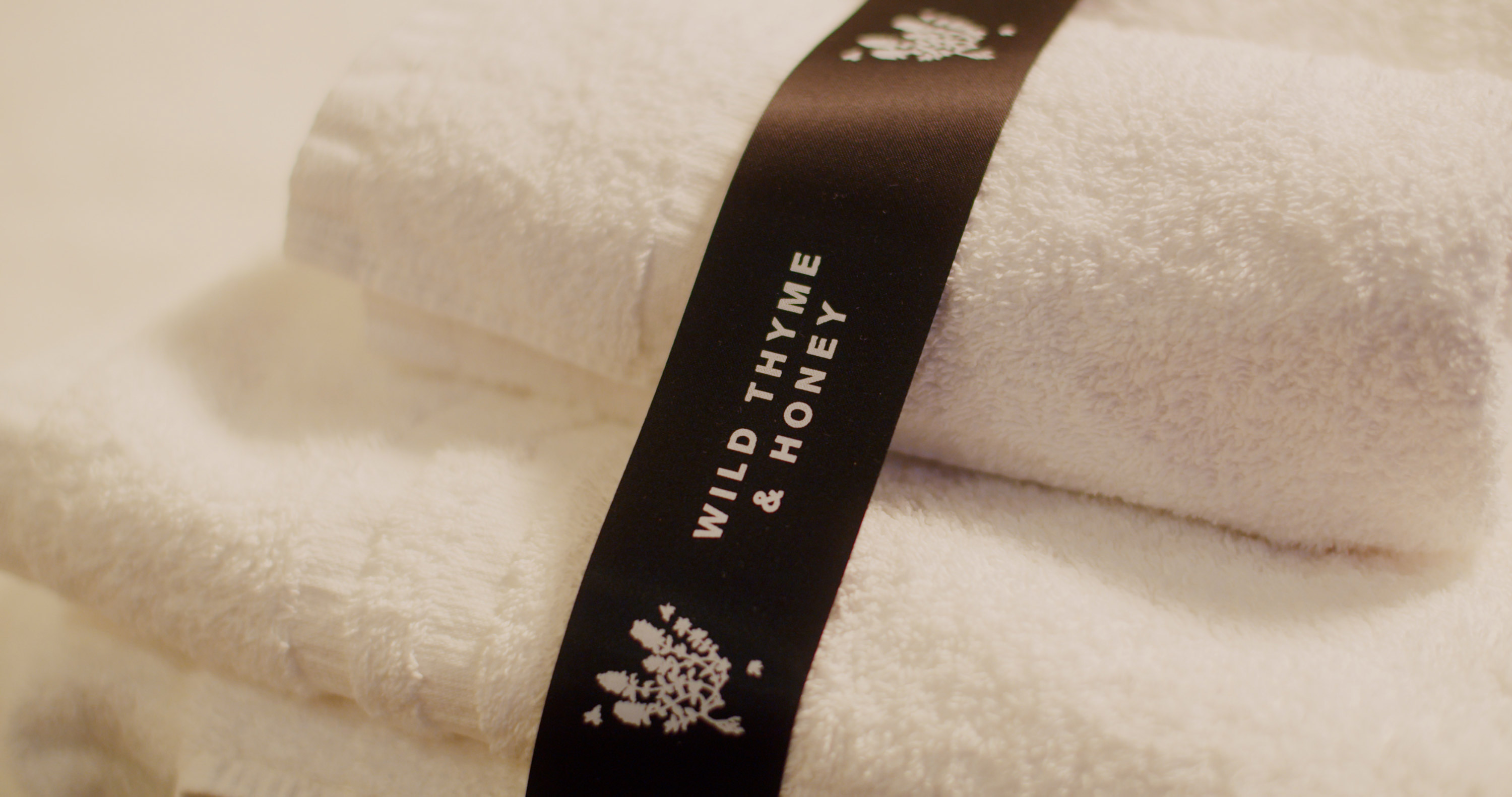 White towels are neatly rolled with a black ribbon wrapped around them. The ribbon features a text label that reads WILD THYME & HONEY along with a simple floral illustration. - Wild Thyme & Honey Hotel in the Cotswolds