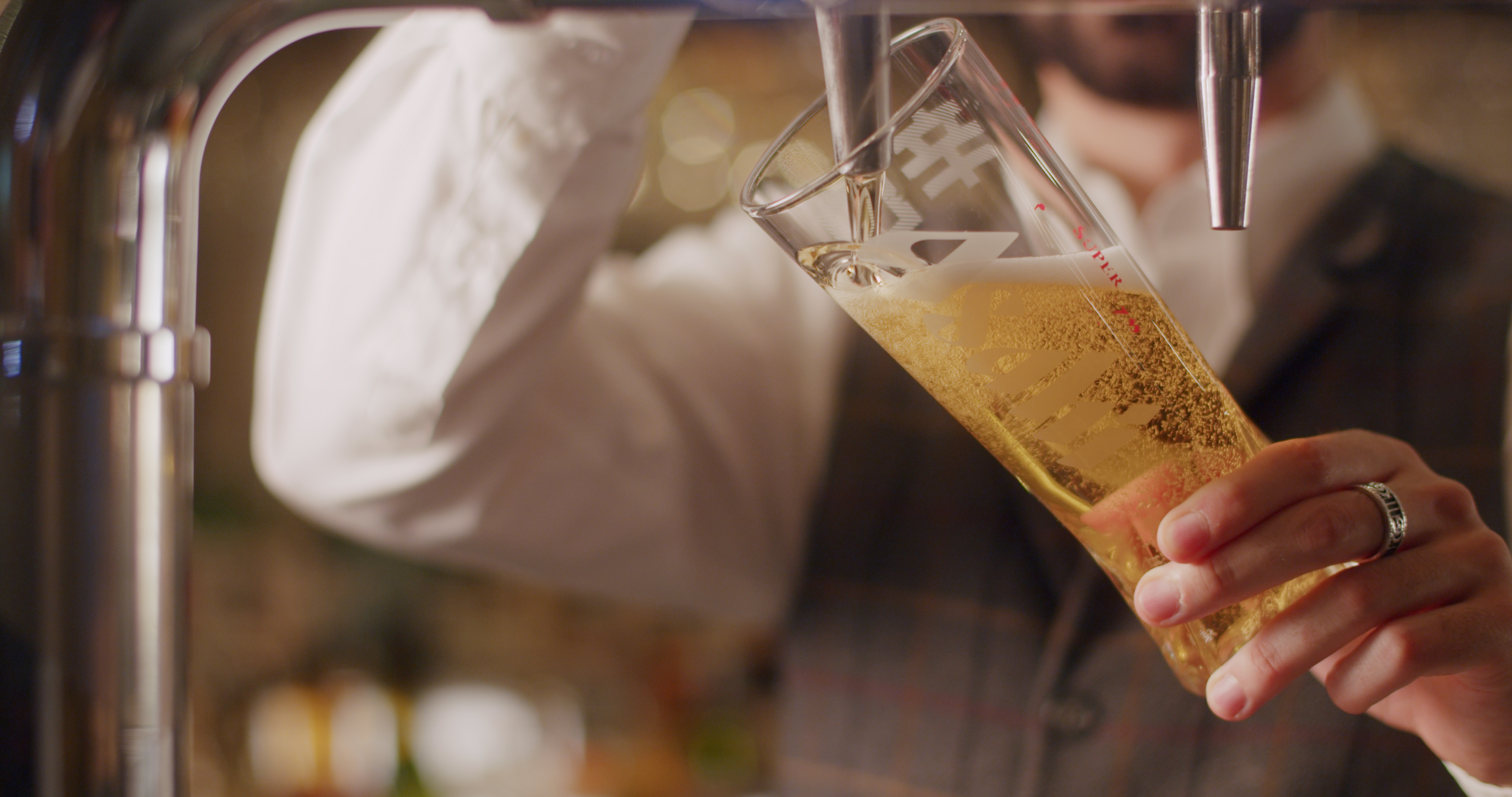 A person fills a tall, narrow glass with draft beer from a tap. Theyre wearing a white shirt and a vest. Bubbles are visible in the light-colored beer. - Wild Thyme & Honey Hotel in the Cotswolds