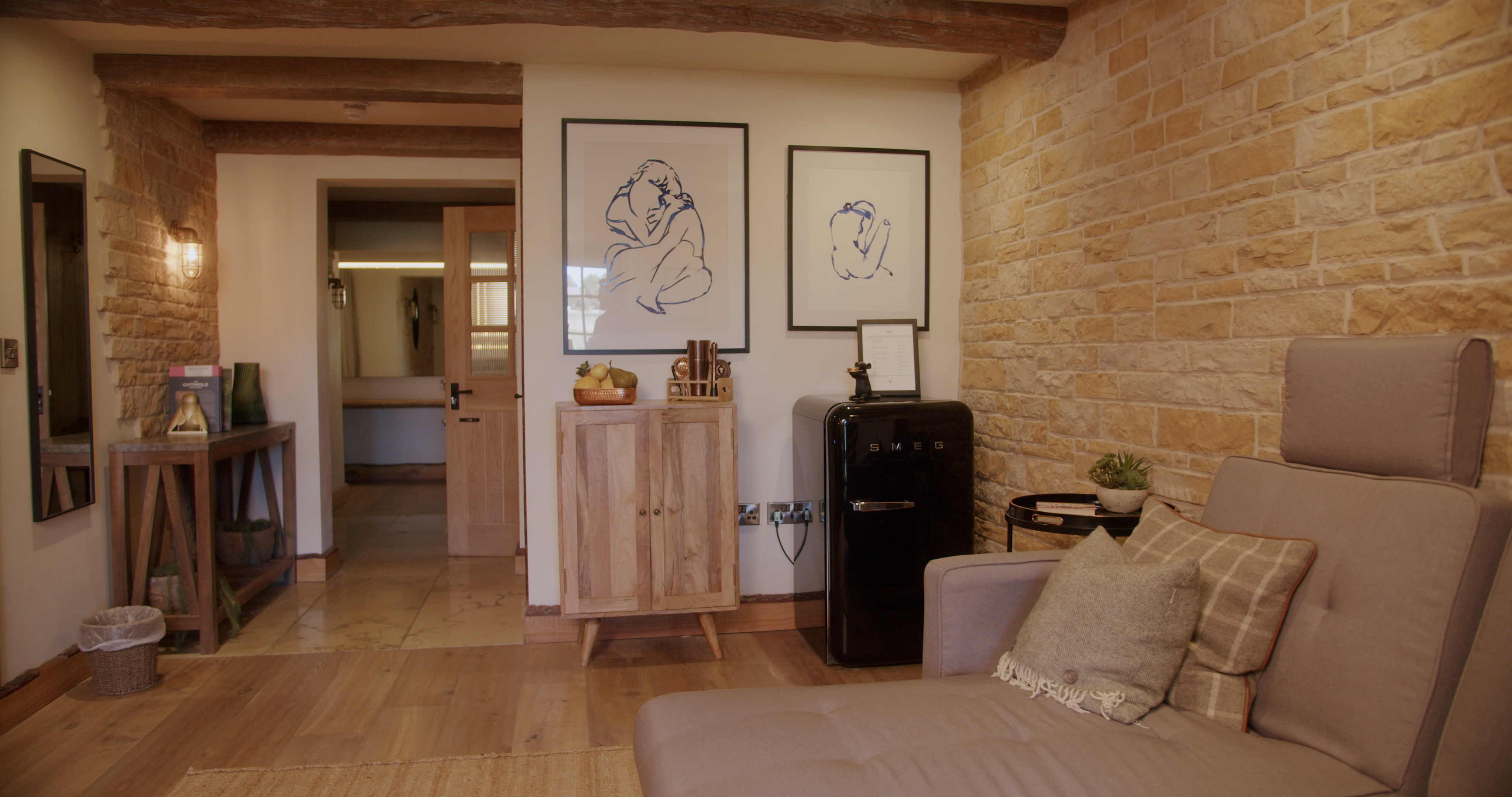 Cozy living room with beige sofa, plaid pillows, and textured walls. A wooden cabinet holds decor beneath line art sketches. Theres a black retro-style fridge, wooden accents, and a hallway view. Subtle lighting and rustic beams complete the ambiance. - Wild Thyme & Honey Hotel in the Cotswolds