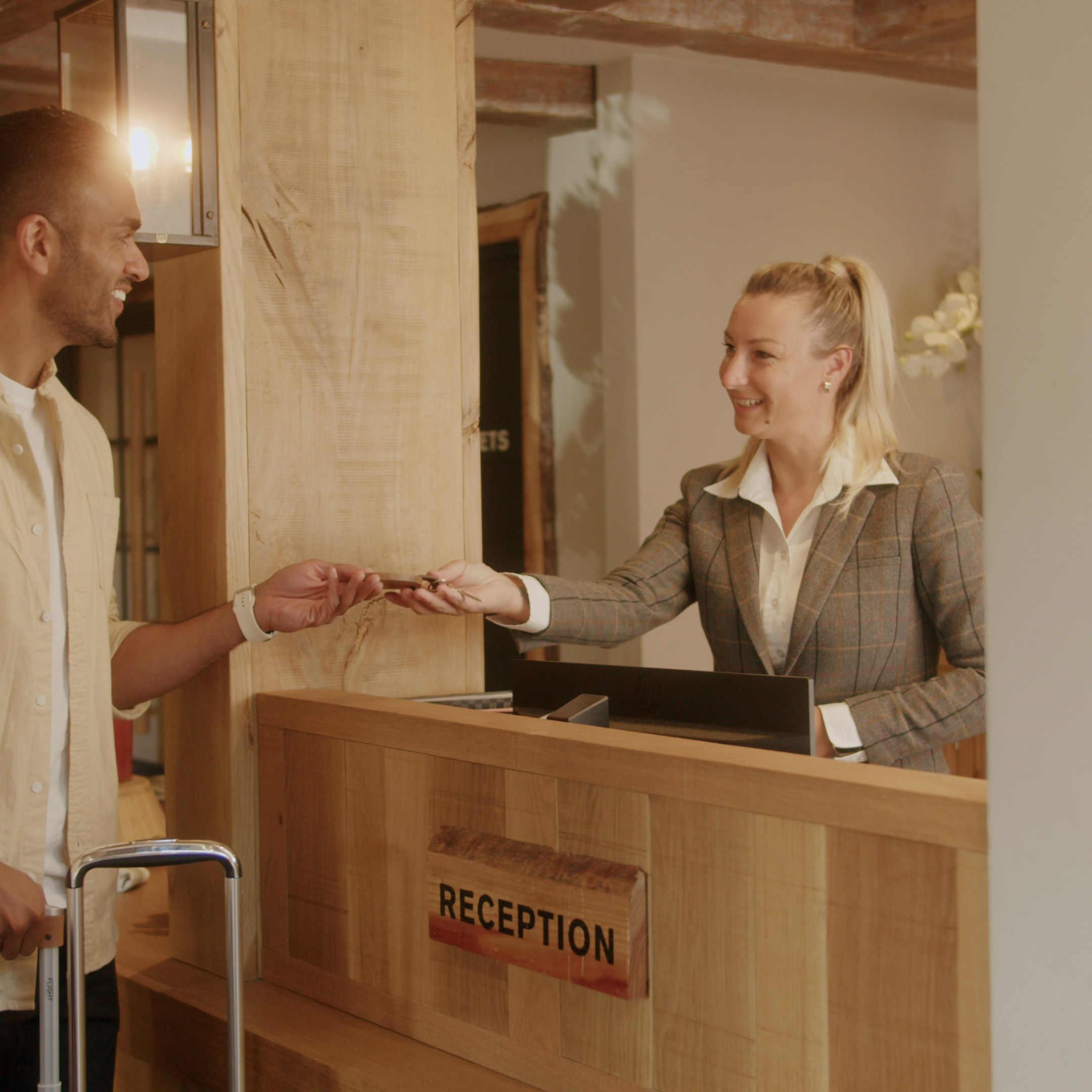 Man handing card to a smiling receptionist at a hotel desk, with a suitcase beside him. - Wild Thyme & Honey Hotel in the Cotswolds