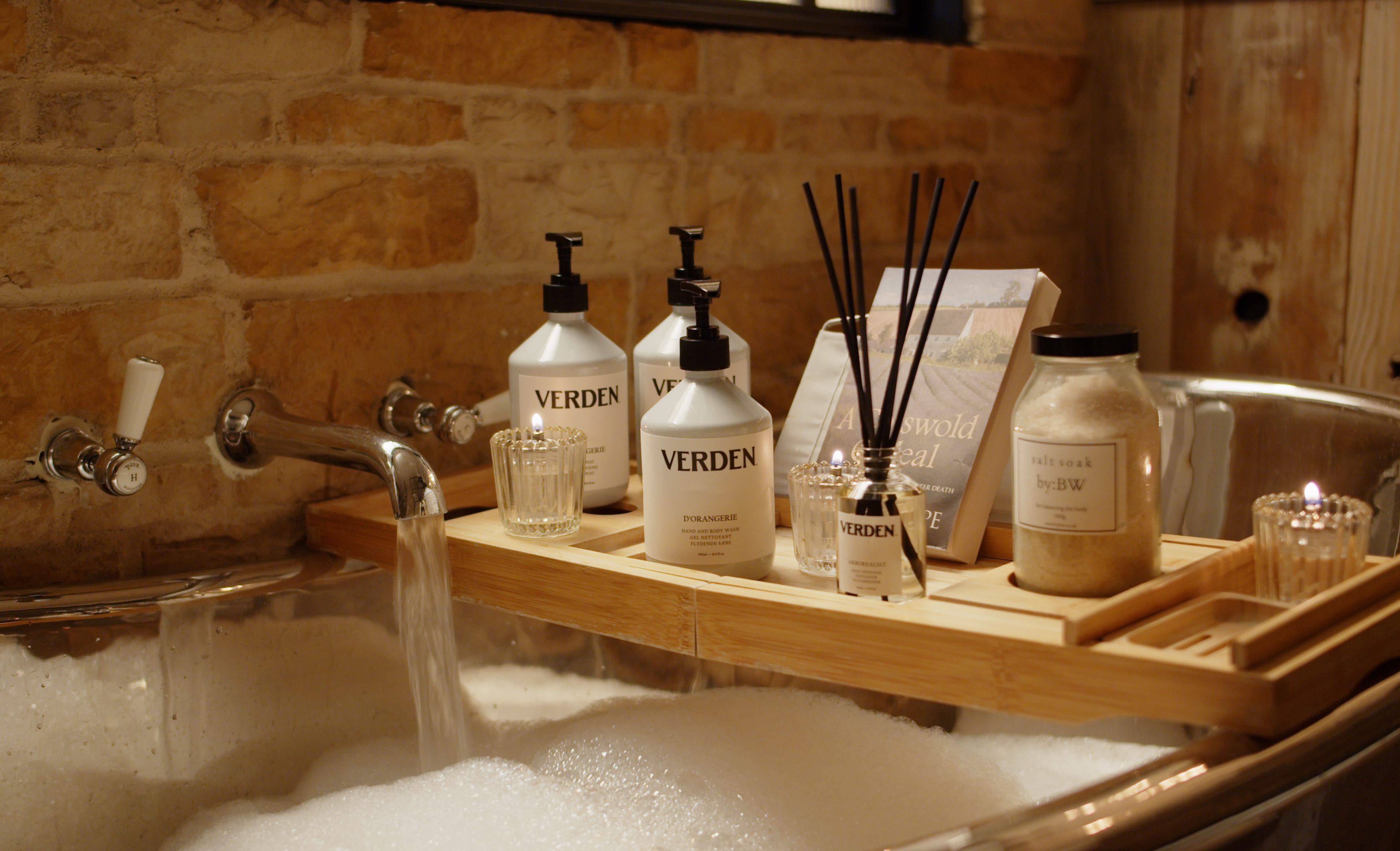 A wooden tray across a bathtub holds bottles and jars of toiletries, along with incense sticks, against a brick wall backdrop. - Wild Thyme & Honey Hotel in the Cotswolds