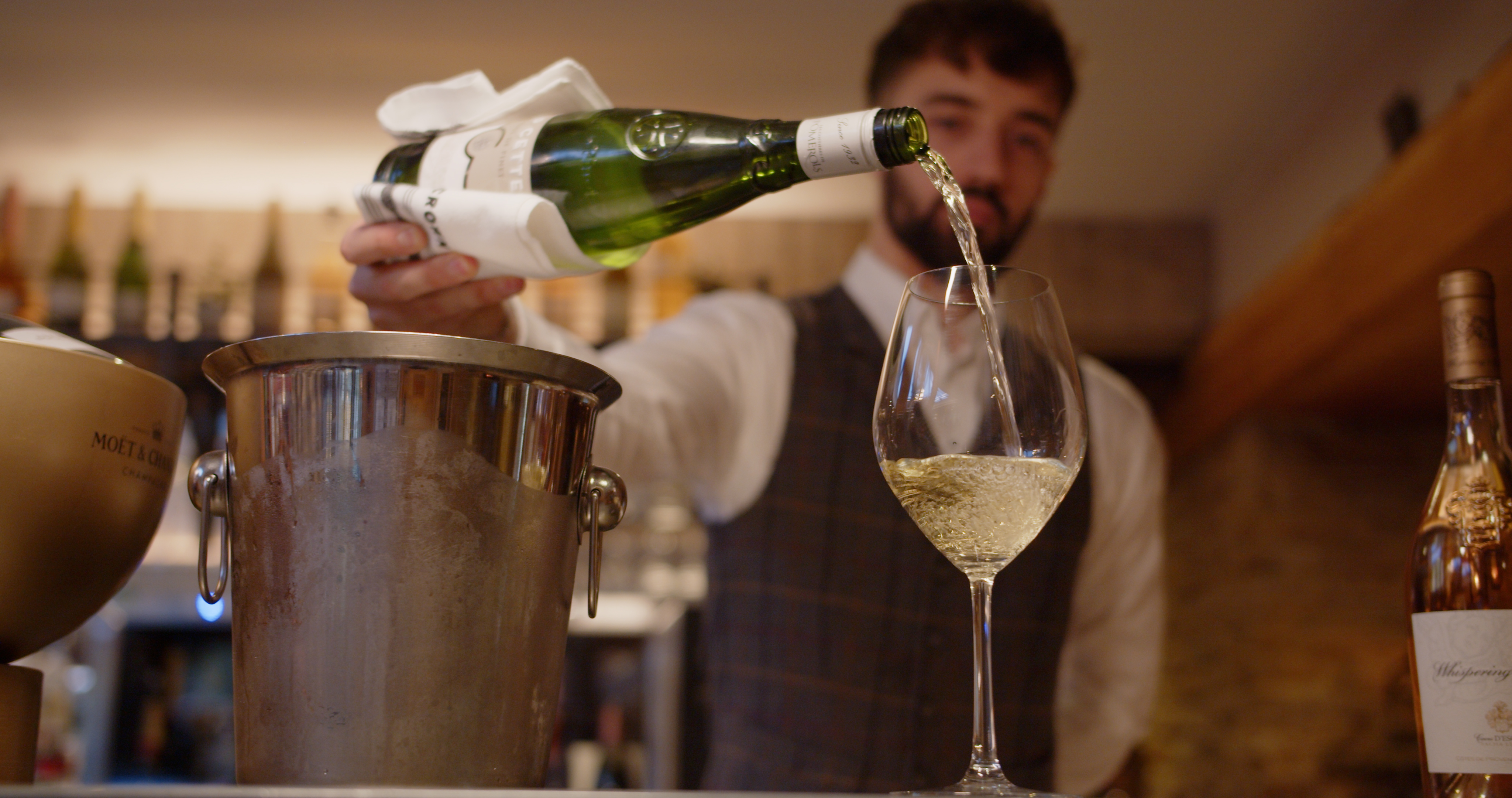 Bartender pouring white wine into a glass, with a bottle cooler and wines in the background. - Wild Thyme & Honey Hotel in the Cotswolds