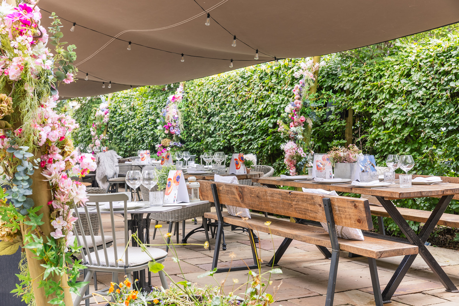 Outdoor restaurant patio with wooden tables, chairs, and floral decorations. Overhead string lights add a cozy ambiance. - Wild Thyme & Honey Hotel in the Cotswolds