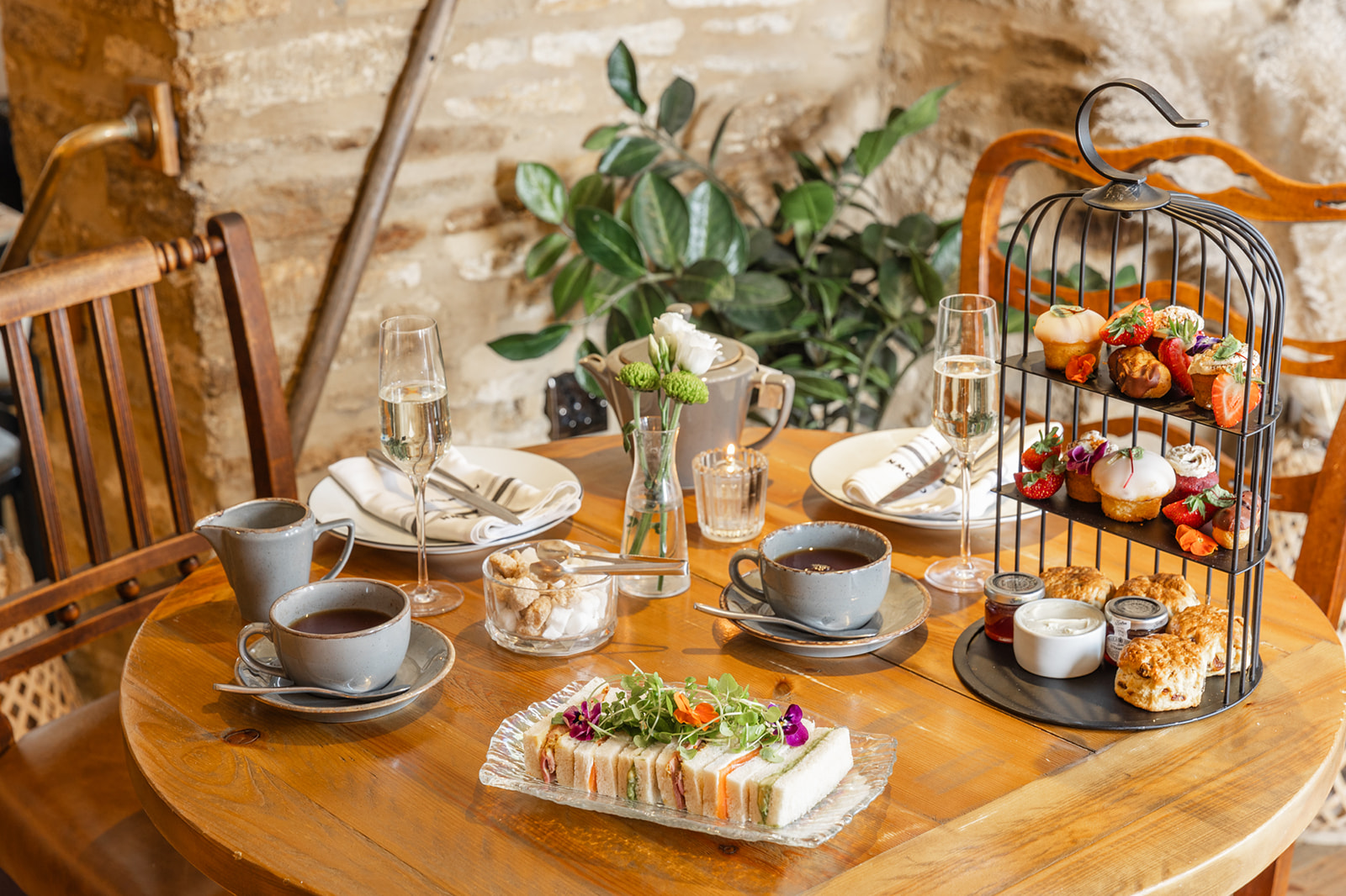A wooden table set for afternoon tea with sandwiches, pastries on a tiered stand, teapots, cups, and glasses of champagne. - Wild Thyme & Honey Hotel in the Cotswolds