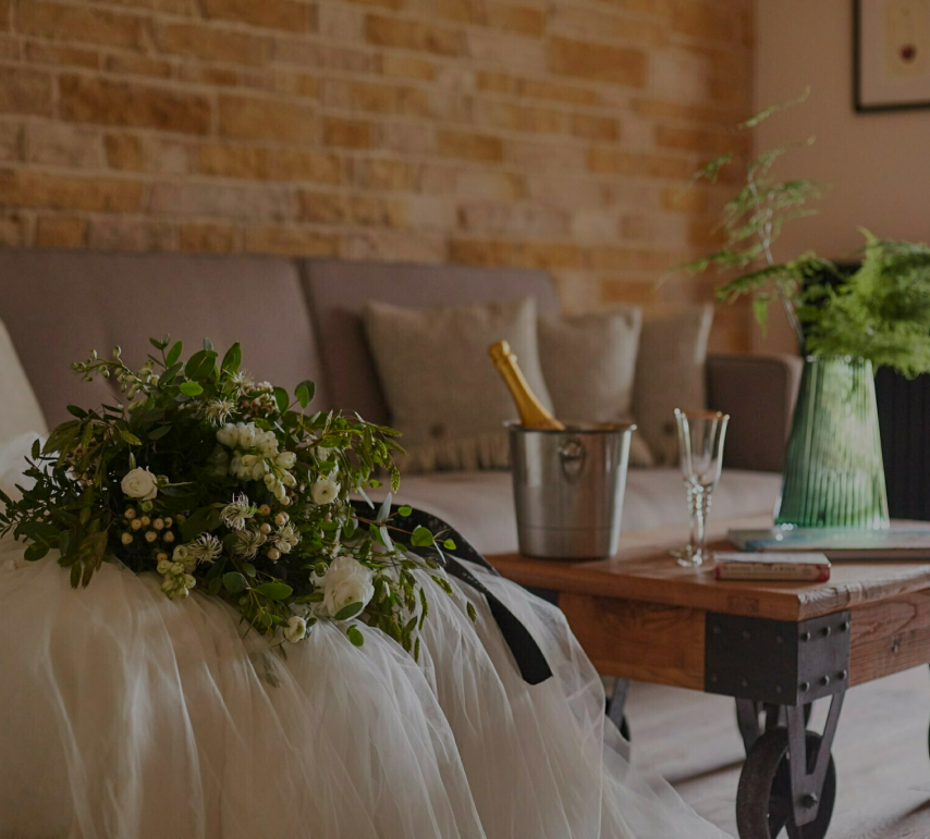 Bridal veil and bouquet on sofa, champagne in an ice bucket, and glasses on a wooden table, with a brick wall in the background. - Wild Thyme & Honey Hotel in the Cotswolds