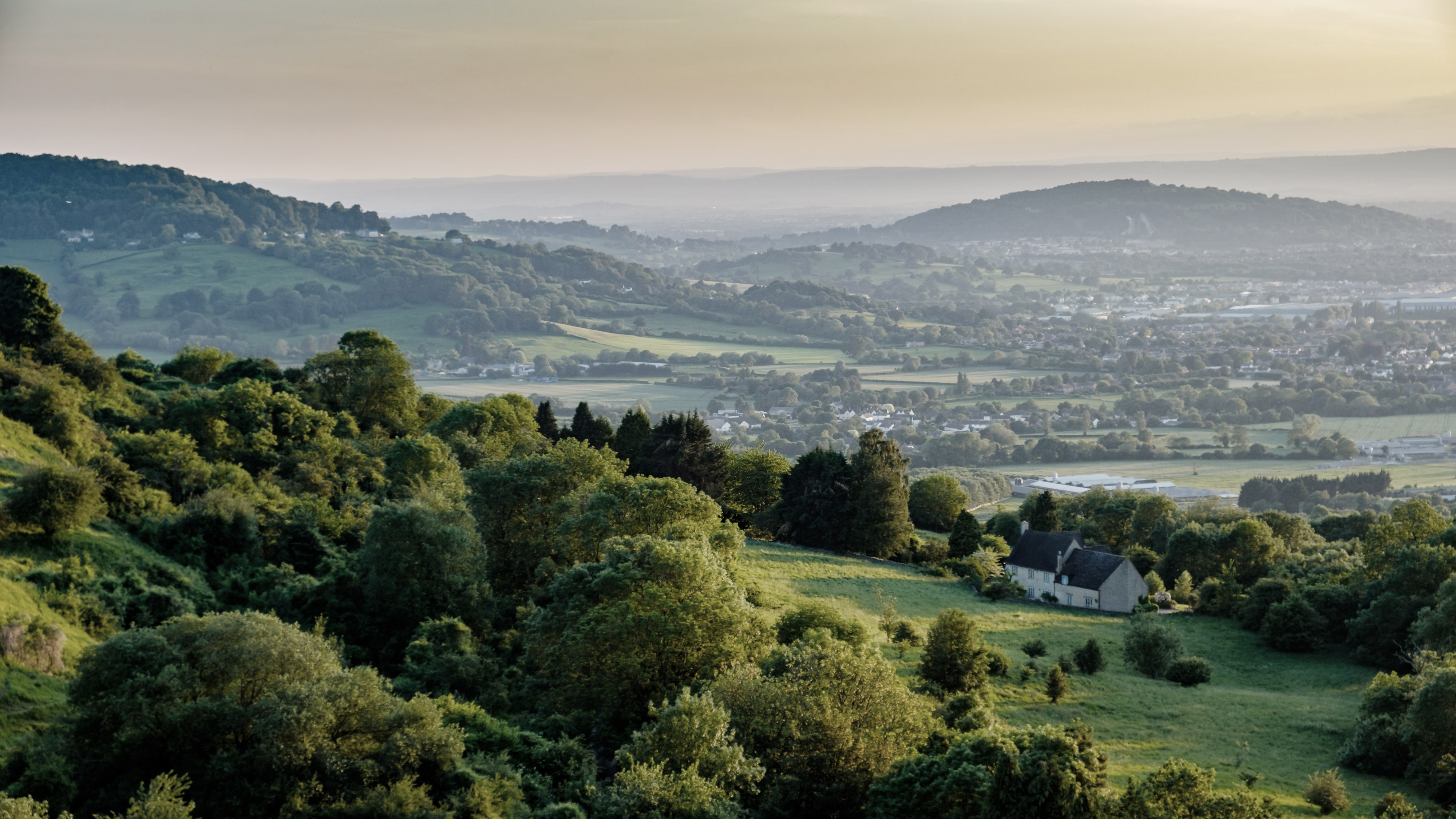 A scenic view of a valley with lush green hills and trees at sunset. A small house is nestled among the greenery in the foreground. Distant hills and a hazy sky are visible in the background. - Wild Thyme & Honey Hotel in the Cotswolds