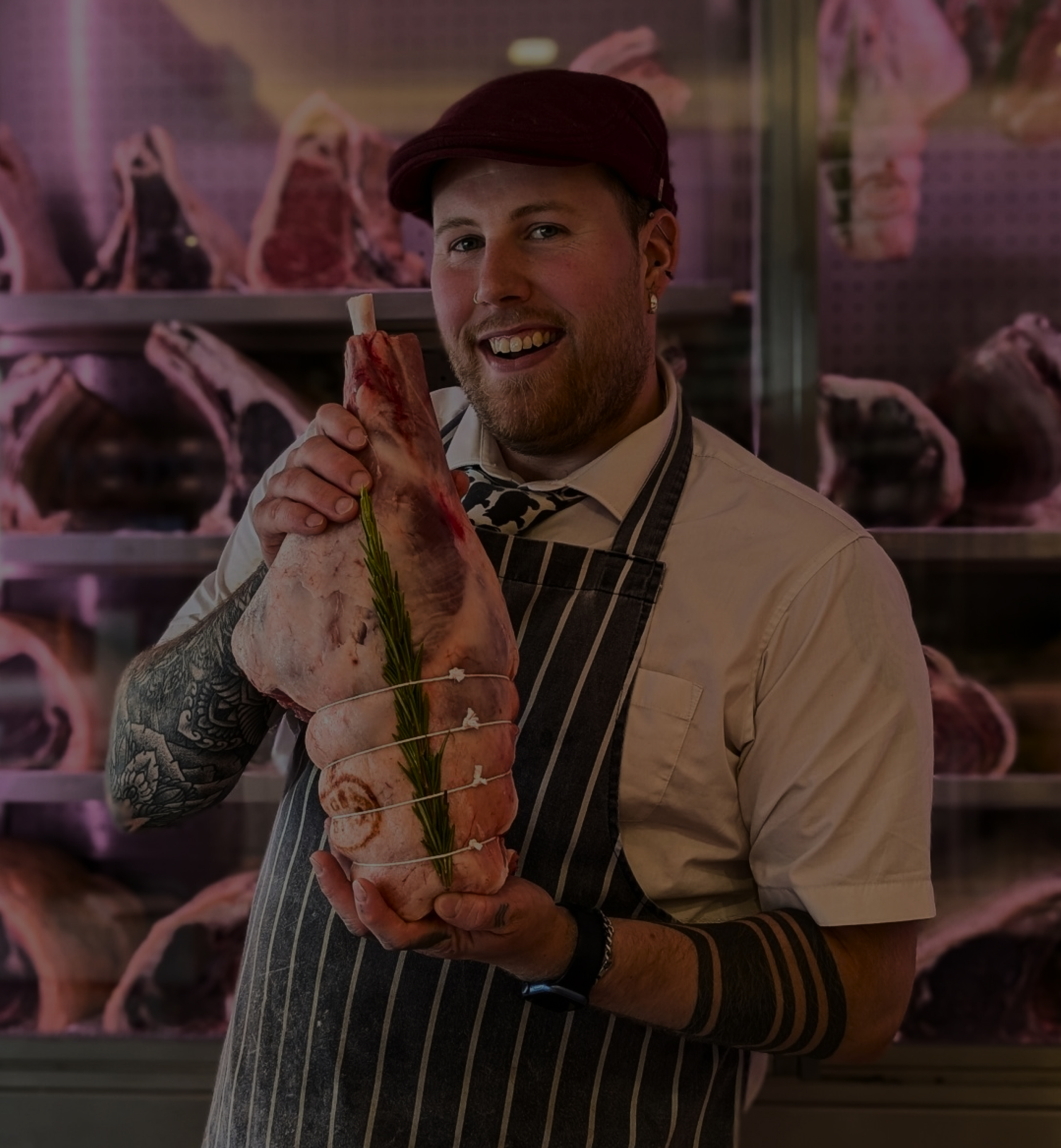 A smiling butcher in a striped apron holds a piece of meat with rosemary. Shelves of meat cuts are visible in the background. - Wild Thyme & Honey Hotel in the Cotswolds