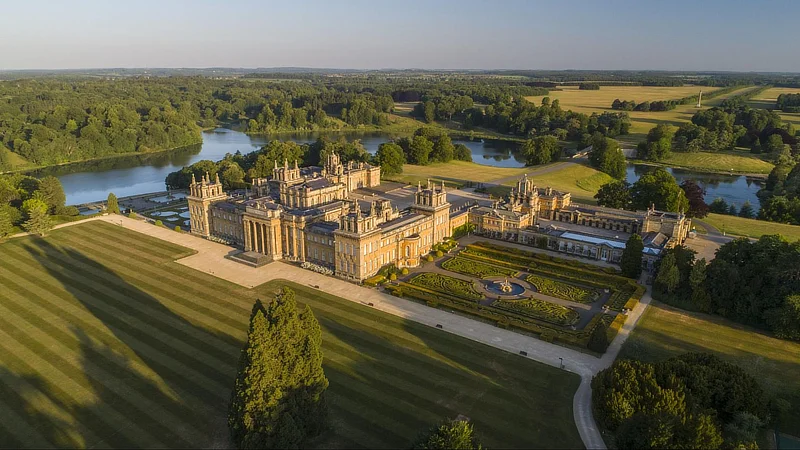 Aerial view of a grand palace surrounded by gardens, trees, and a lake under a clear blue sky. - Wild Thyme & Honey Hotel in the Cotswolds