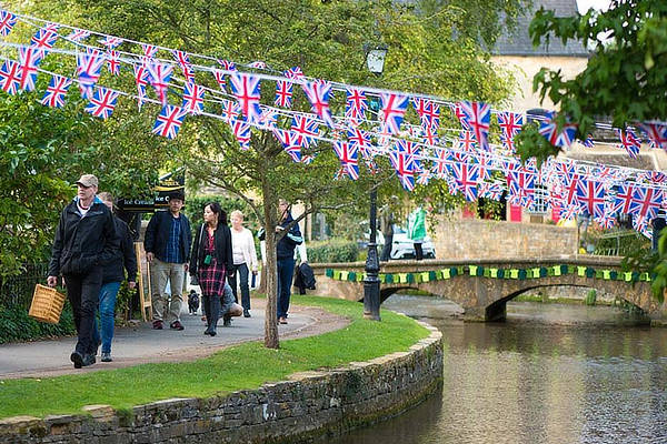 People walk along a riverside path decorated with Union Jack bunting. A stone bridge and lush trees are in the background. - Wild Thyme & Honey Hotel in the Cotswolds