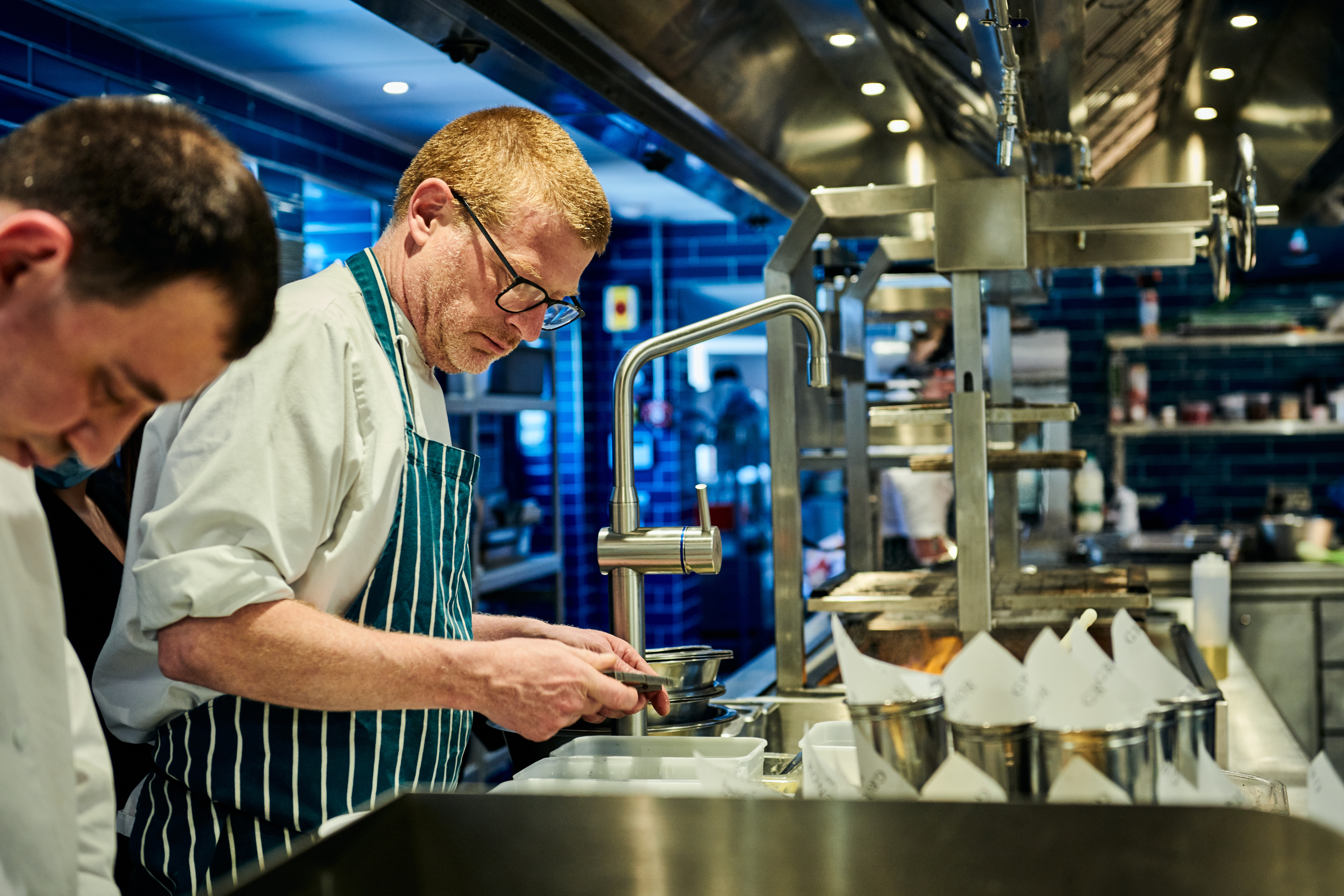 Two chefs working in a bustling kitchen; one focuses on plating a dish while wearing a striped apron. - Wild Thyme & Honey Hotel in the Cotswolds