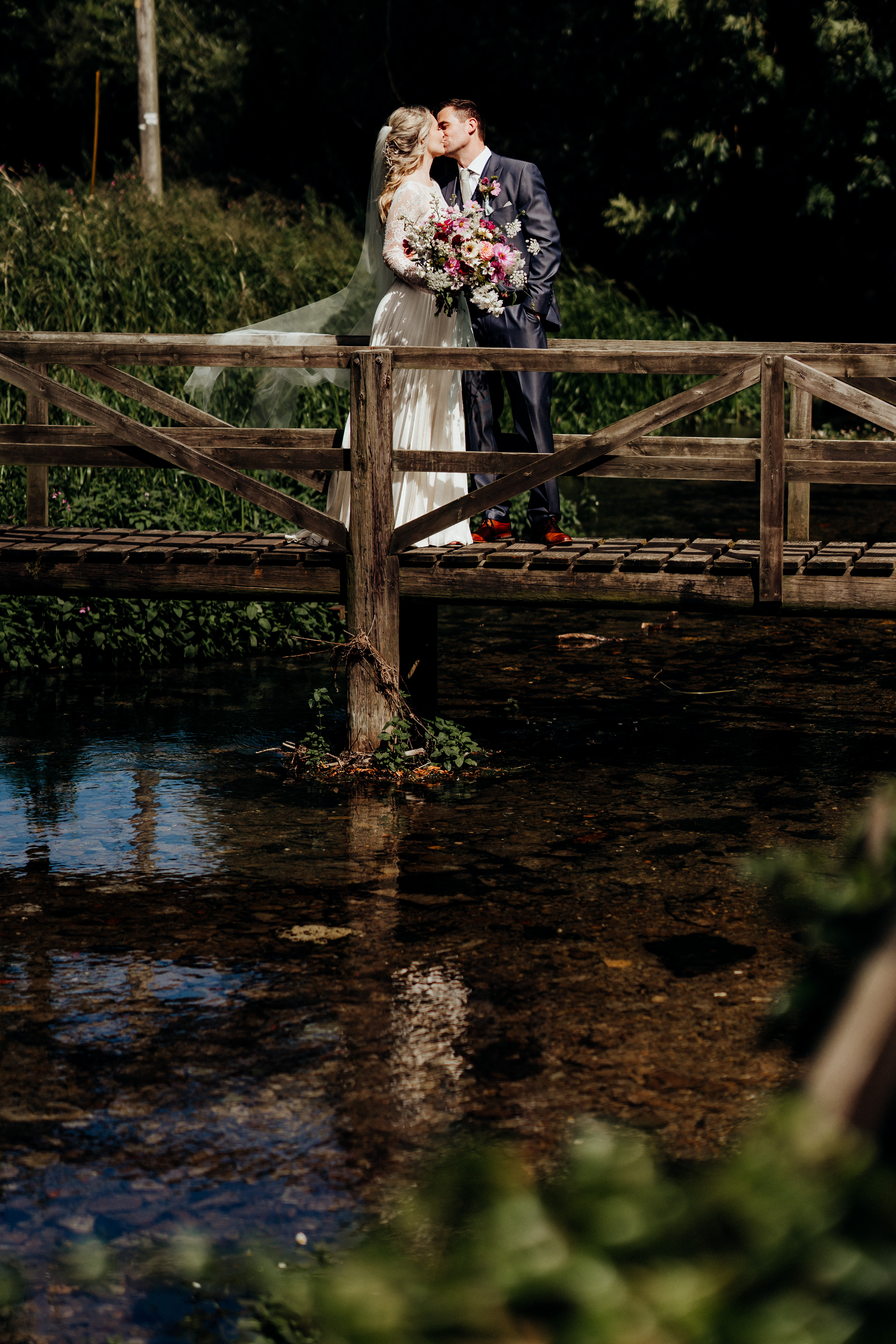 A bride and groom kiss on a wooden bridge over a serene stream, surrounded by lush greenery and holding a bouquet of flowers. - Wild Thyme & Honey Hotel in the Cotswolds