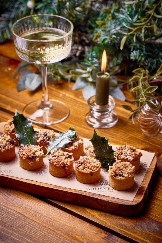 Mini tarts with holly garnish on a wooden board, next to a glass of white wine and a lit candle, with greenery in the background. - Wild Thyme & Honey Hotel in the Cotswolds