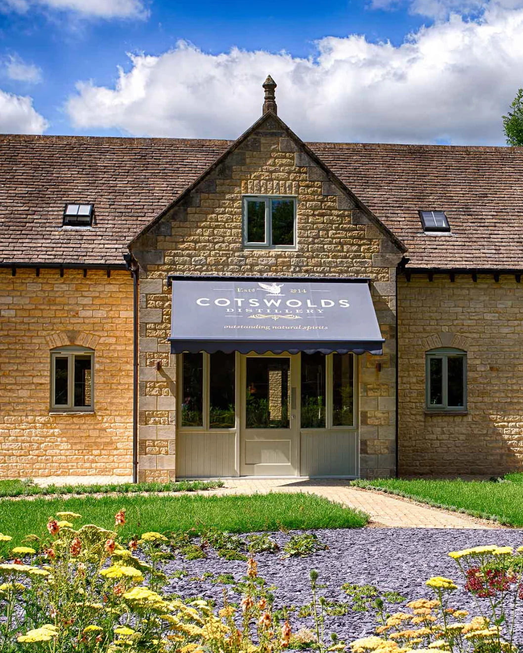 Stone building with a blue awning labeled Cotswolds, set against a cloudy blue sky, surrounded by green grass and flowers. - Wild Thyme & Honey Hotel in the Cotswolds