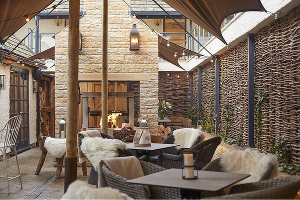 Cozy outdoor patio with wicker chairs, furry throws, and a stone fireplace under a canopy. Rustic decor and warm lighting. - Wild Thyme & Honey Hotel in the Cotswolds