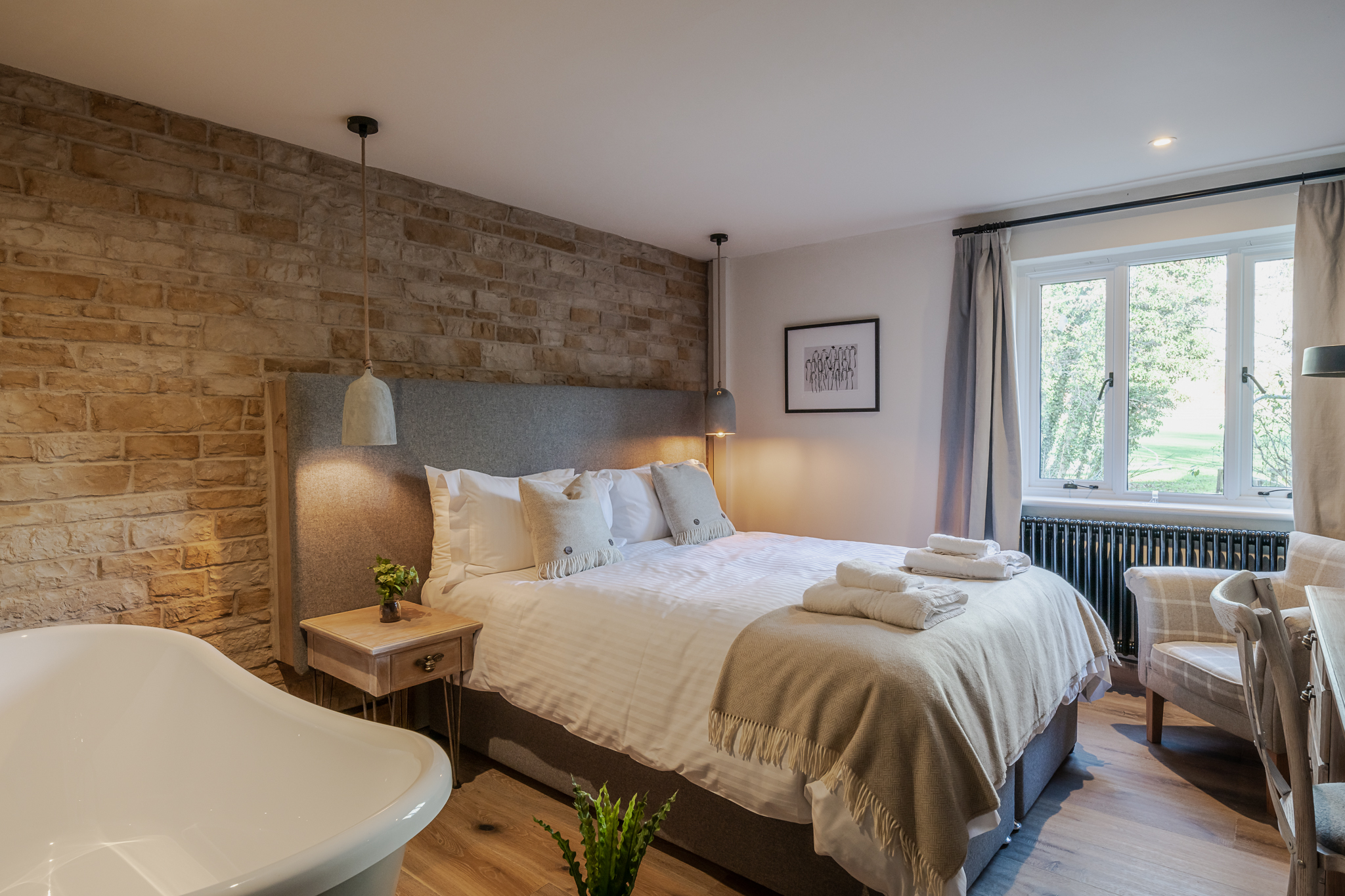 Cozy bedroom with a stone accent wall, double bed, two pendant lights, armchair, window view, and a standalone bathtub. - Wild Thyme & Honey Hotel in the Cotswolds