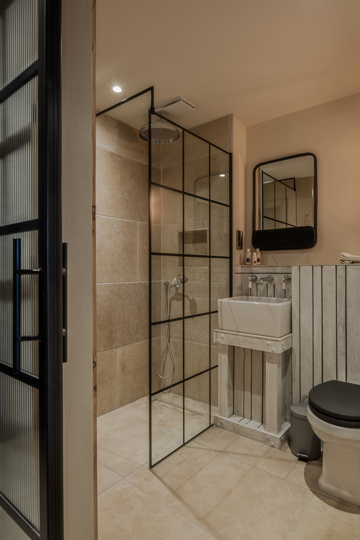 Modern bathroom with beige tiles, glass shower partition, rectangular sink with mirror, and toilet with black seat. - Wild Thyme & Honey Hotel in the Cotswolds