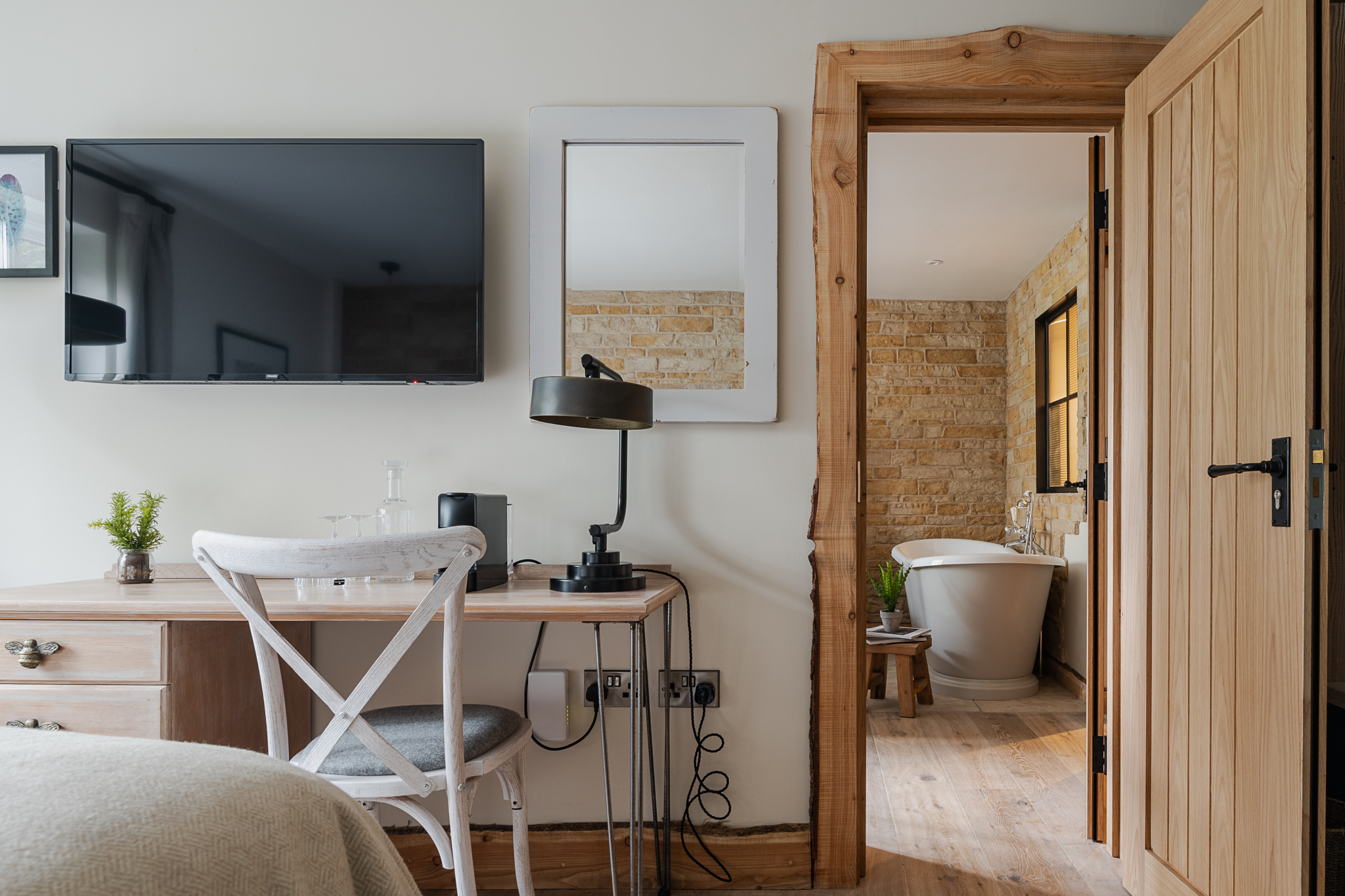 A cozy bedroom features a desk with a lamp, TV on the wall, open door to a bathroom with a bathtub, and beige tones throughout. - Wild Thyme & Honey Hotel in the Cotswolds