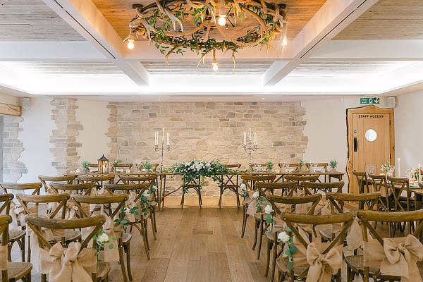 Rustic wedding ceremony setup with wooden chairs, floral decorations, and a stone wall backdrop under a chandelier with greenery. - Wild Thyme & Honey Hotel in the Cotswolds