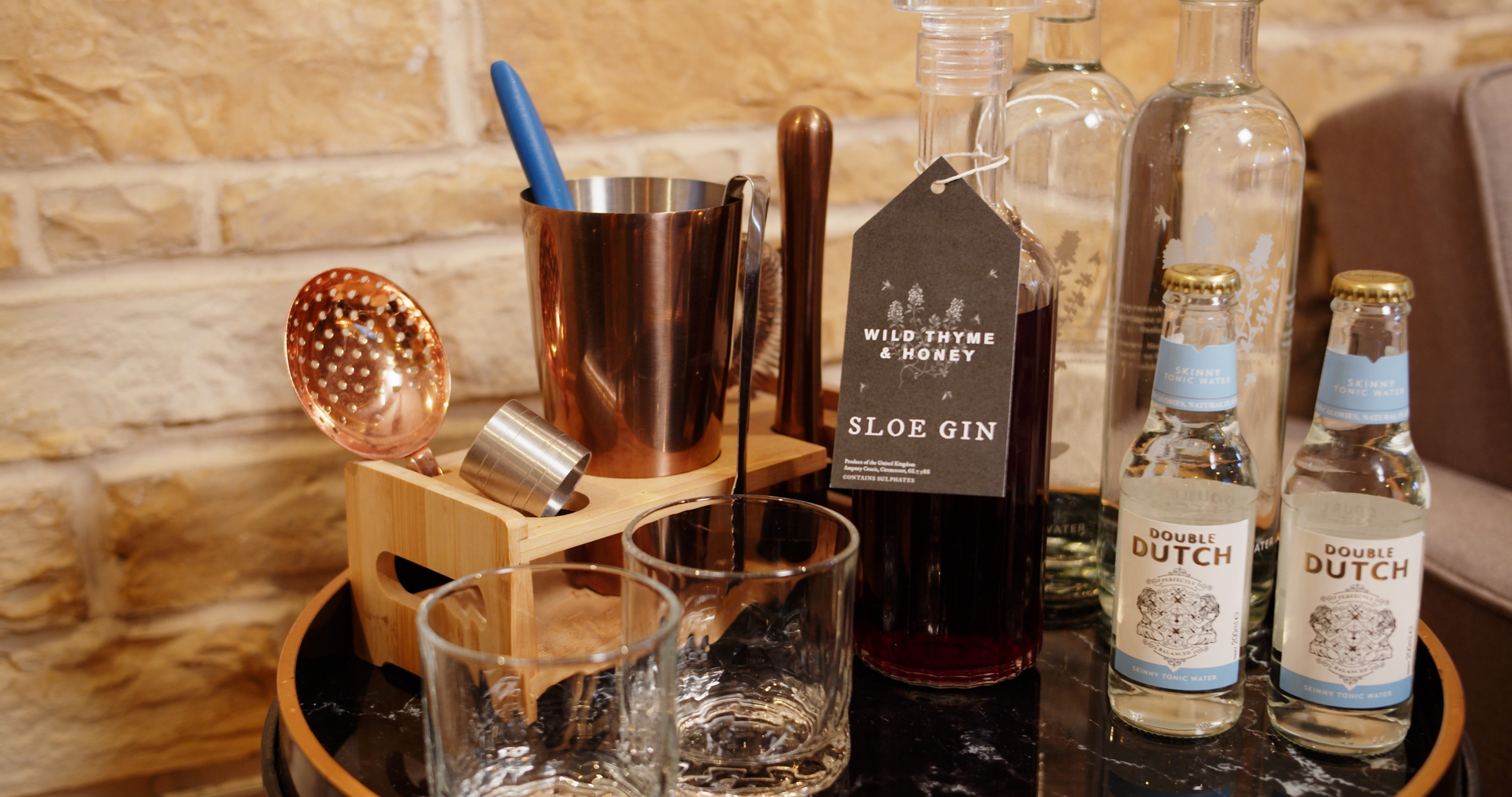 A bar setup with glasses, copper utensils, sloe gin, and tonic water bottles on a wooden countertop in front of a brick wall. - Wild Thyme & Honey Hotel in the Cotswolds