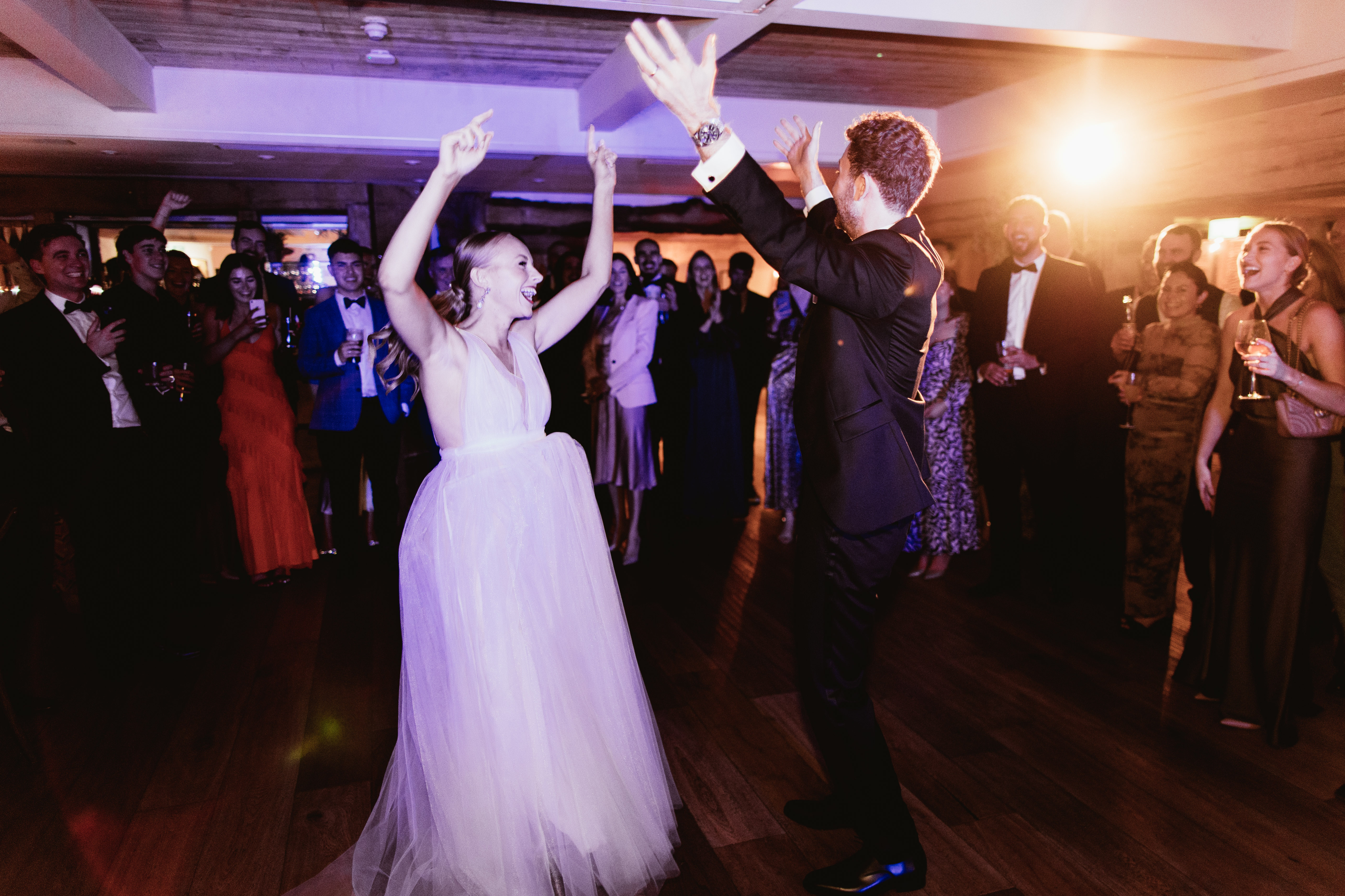 A bride and groom dancing energetically at a wedding reception while guests cheer and celebrate around them. - Wild Thyme & Honey Hotel in the Cotswolds