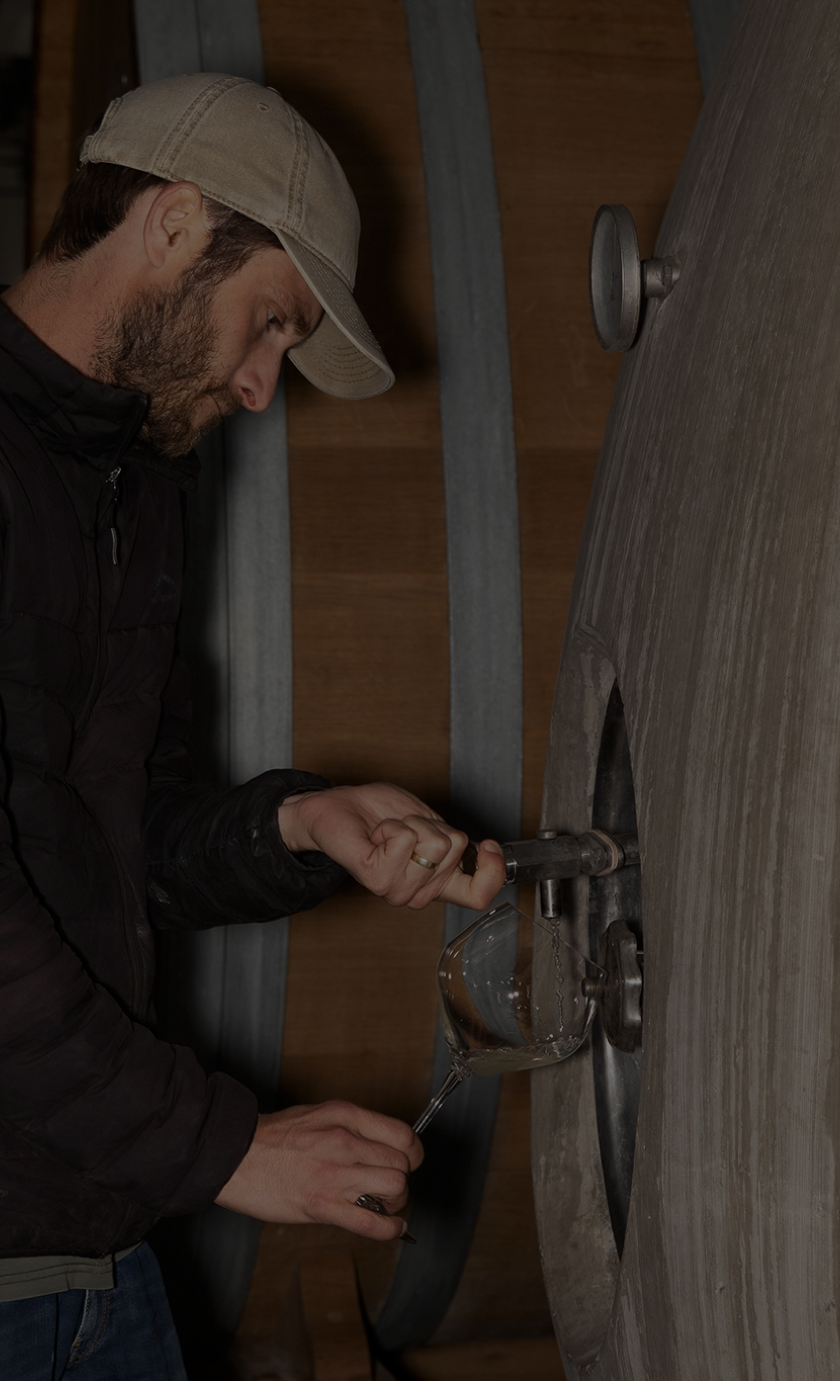 Person in a cap and jacket draws wine from a cask into a glass in a dimly lit cellar. - Wild Thyme & Honey Hotel in the Cotswolds