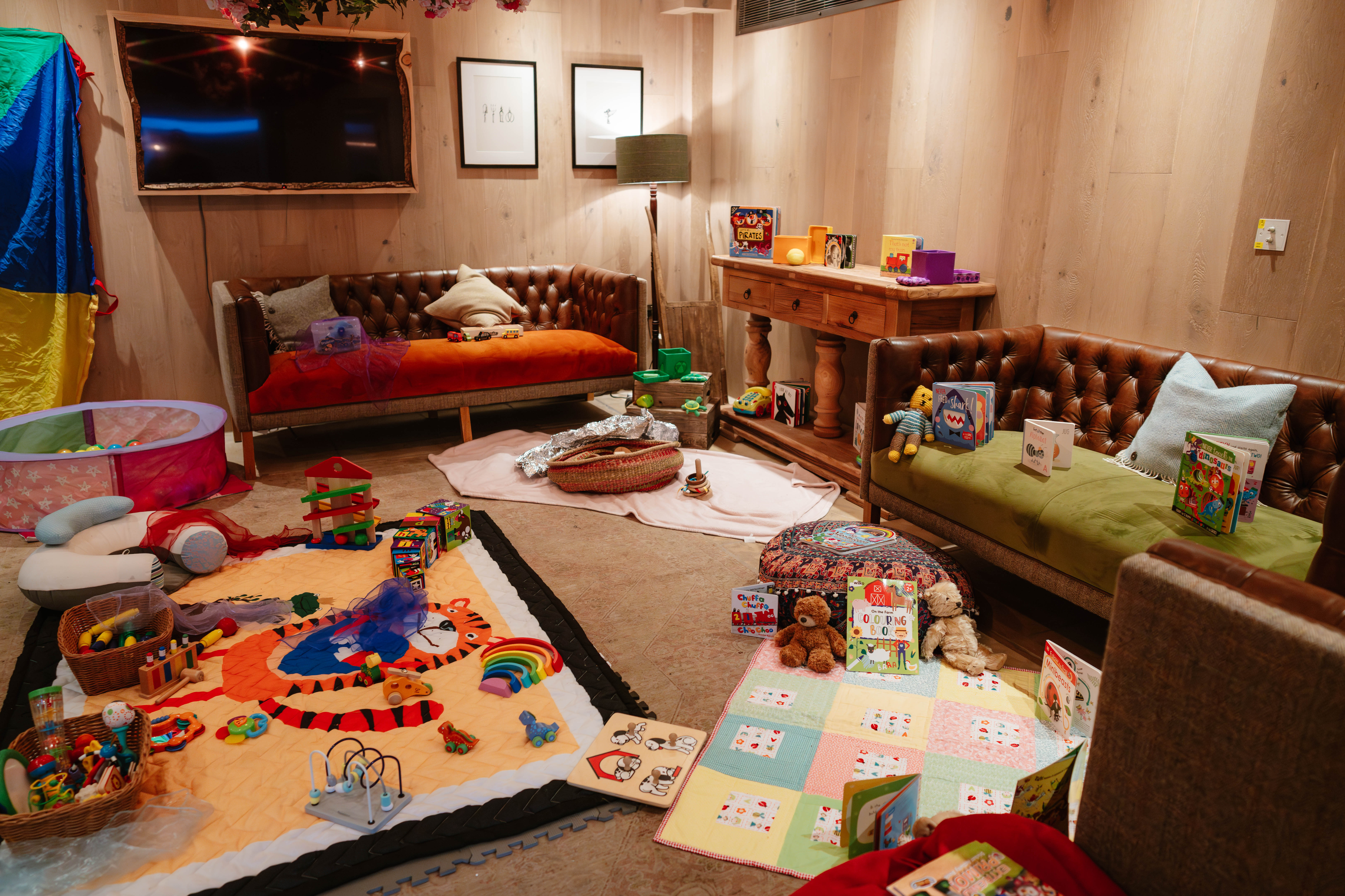 Cozy playroom with toy-filled area, brown leather sofas, a TV, a wooden table, and colorful play mats on the floor. - Wild Thyme & Honey Hotel in the Cotswolds