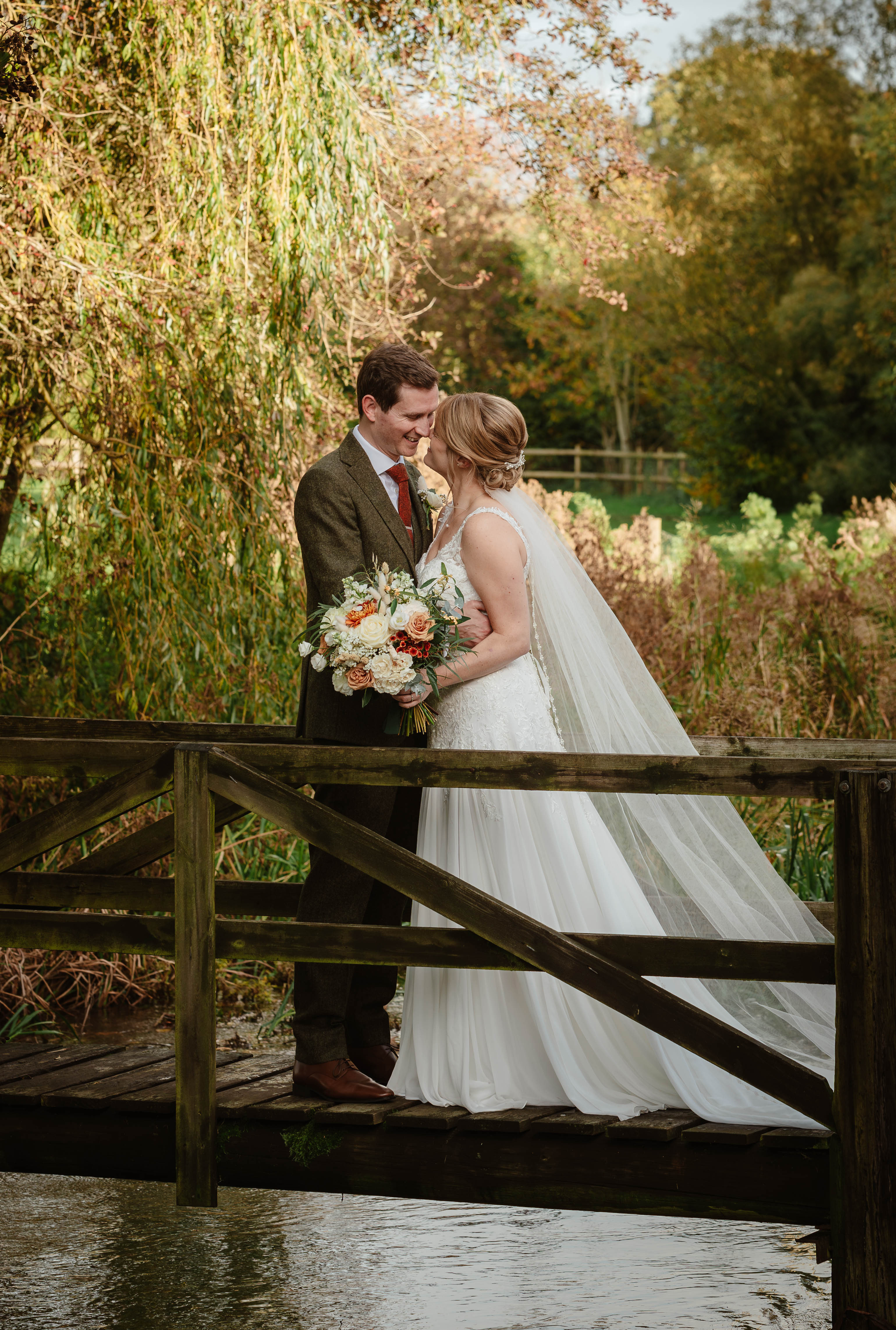 Bride and groom standing on a wooden bridge, embracing with a bouquet, surrounded by lush greenery. - Wild Thyme & Honey Hotel in the Cotswolds