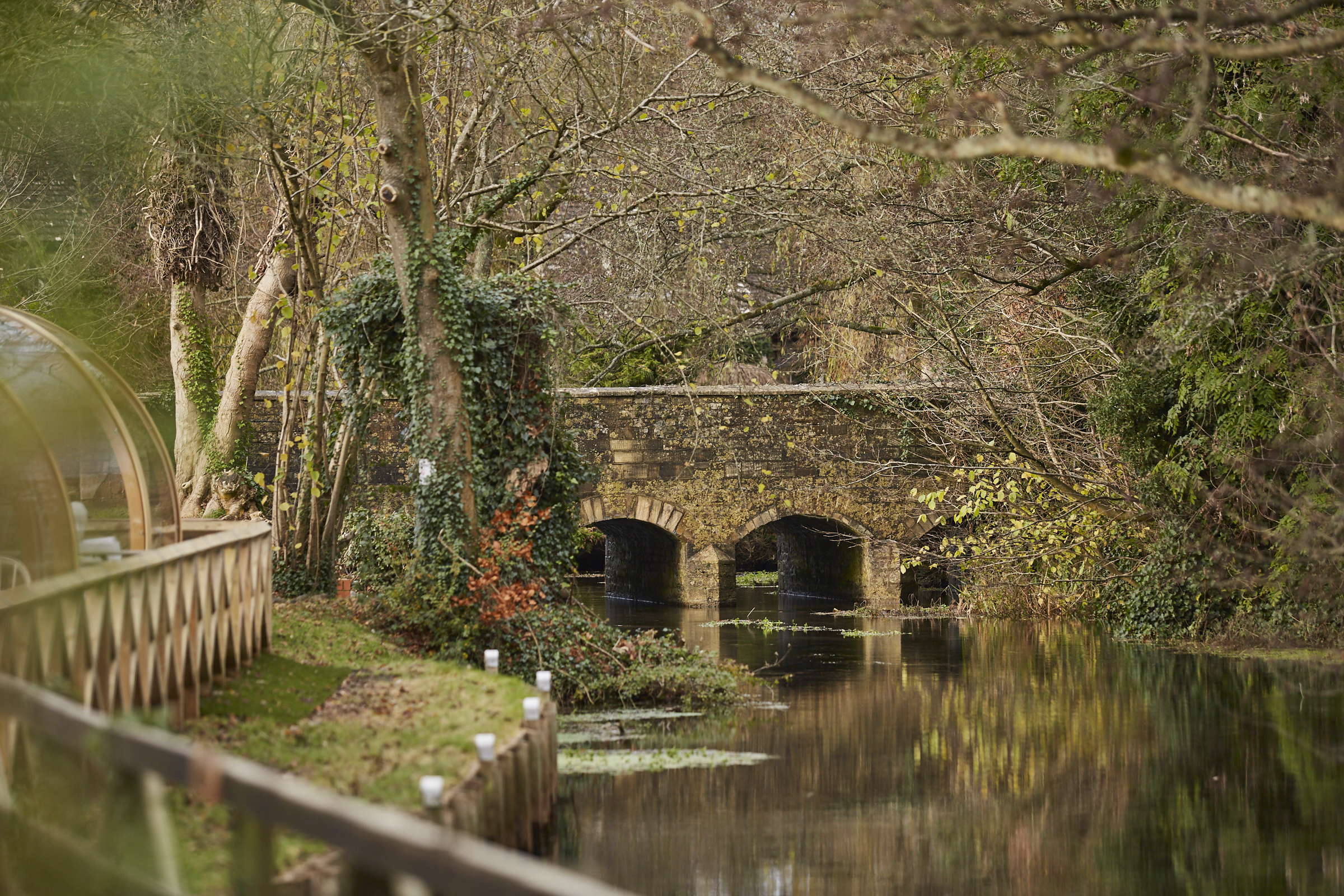 A stone bridge with three arches spans over a calm river, surrounded by trees with sparse winter foliage. - Wild Thyme & Honey Hotel in the Cotswolds
