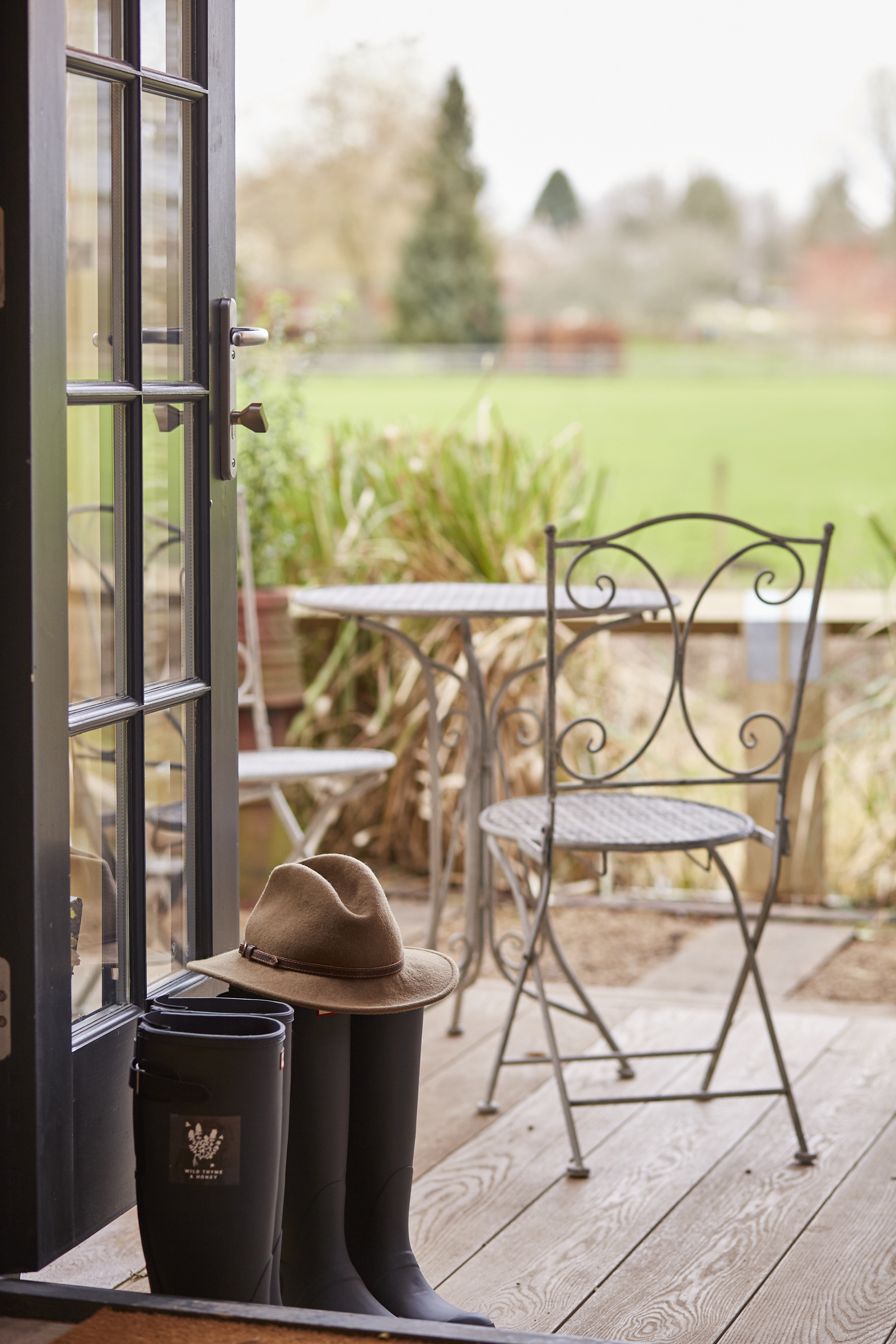 A patio view with a chair, table, hat on a bucket, and boots by an open door, overlooking a grassy field and trees. - Wild Thyme & Honey Hotel in the Cotswolds