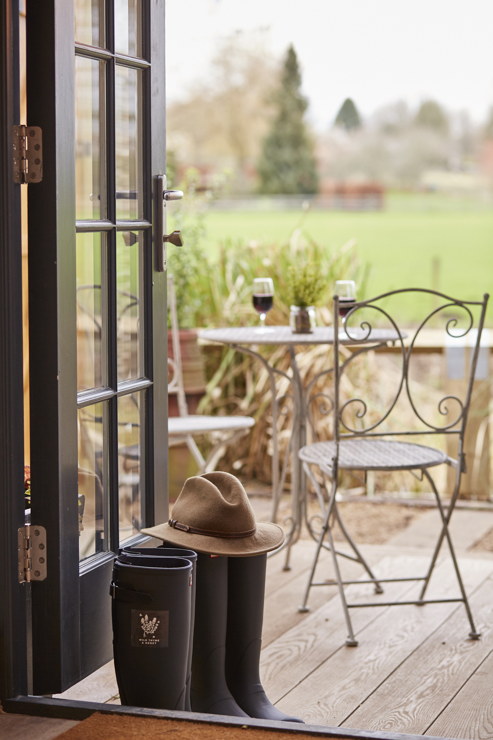 An open door leads to a patio with a table and chairs. A pair of boots and a hat rest by the door. Lush greenery is visible. - Wild Thyme & Honey Hotel in the Cotswolds