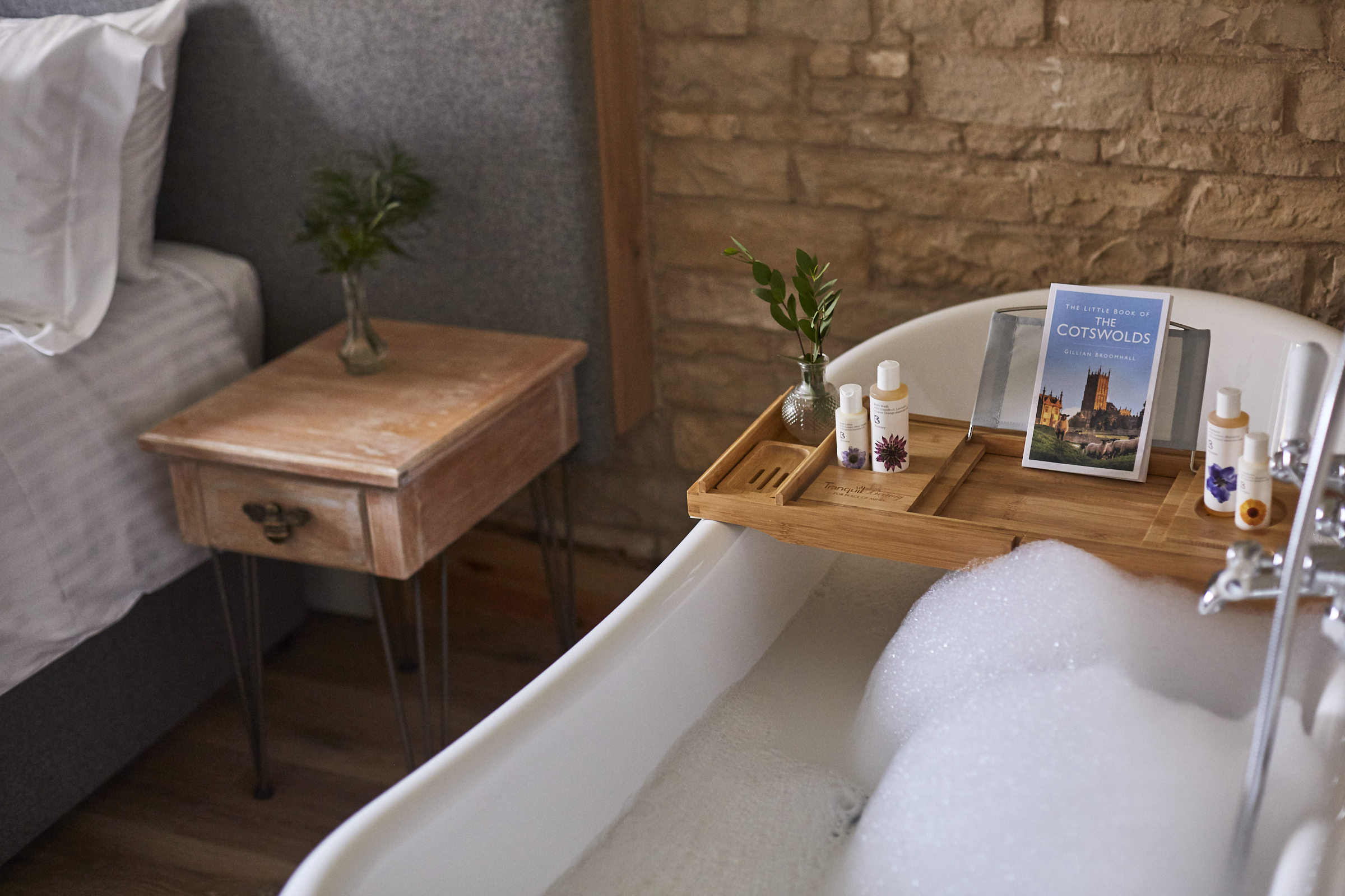 A cozy bathroom scene with a bubble-filled bathtub, wooden tray holding toiletries, a book, and a potted plant nearby. - Wild Thyme & Honey Hotel in the Cotswolds