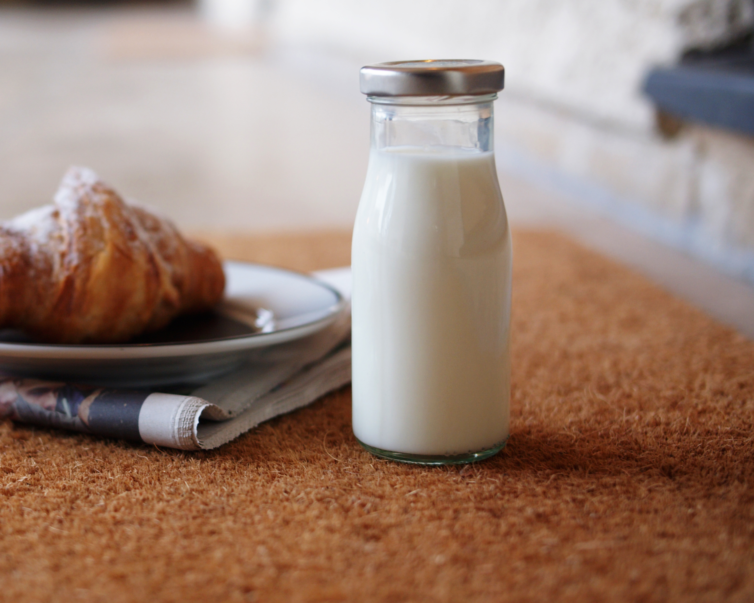 A glass bottle of milk next to a plate with a croissant and a newspaper on a brown mat. - Wild Thyme & Honey Hotel in the Cotswolds