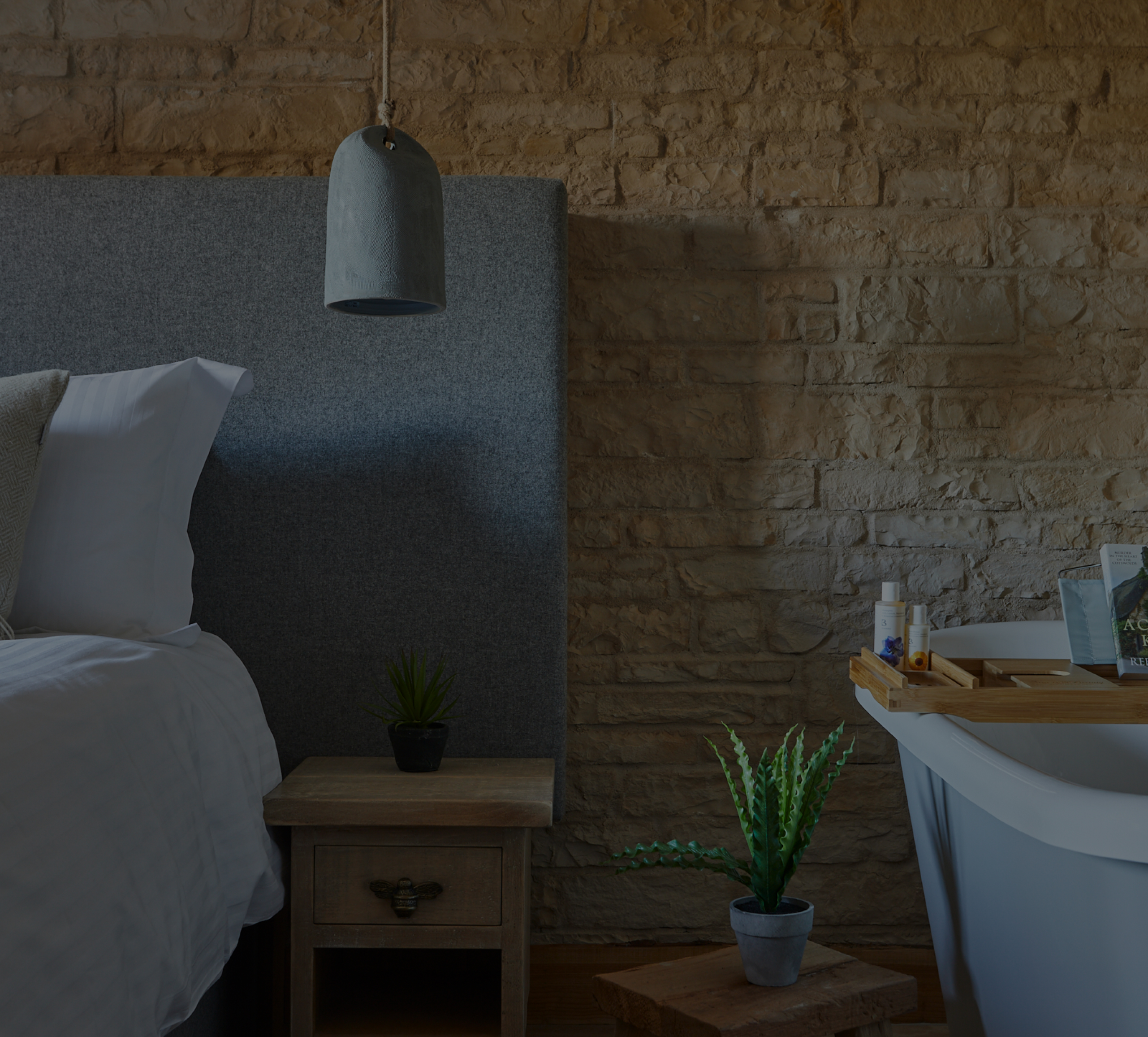 Cozy bedroom with a gray headboard and rustic stone wall. A potted plant sits on a side table near a hanging lamp and bathtub. - Wild Thyme & Honey Hotel in the Cotswolds