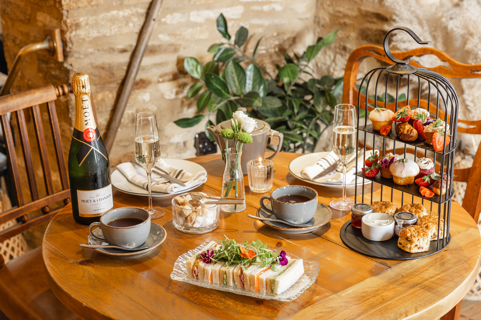 Cozy table setting with tea, champagne, sandwiches, pastries on a tiered stand, and a rustic background with greenery. - Wild Thyme & Honey Hotel in the Cotswolds