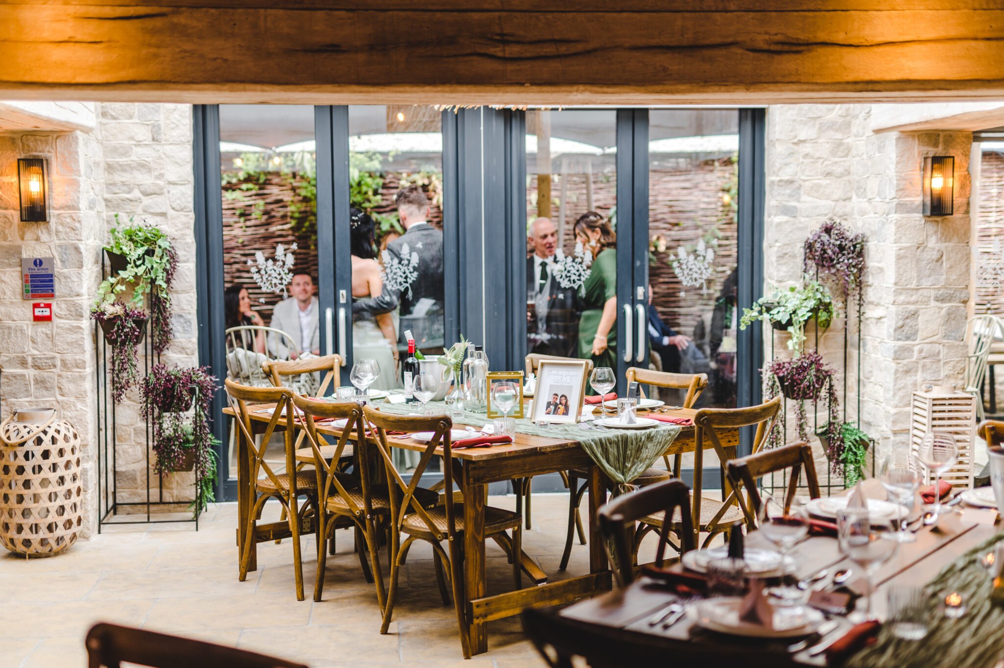 Elegant dining area with wooden chairs and tables, decorative plants, and framed photos. Glass doors reveal people outside. - Wild Thyme & Honey Hotel in the Cotswolds