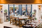 Elegant dining area with wooden chairs and tables, decorative plants, and framed photos. Glass doors reveal people outside. - Wild Thyme & Honey Hotel in the Cotswolds