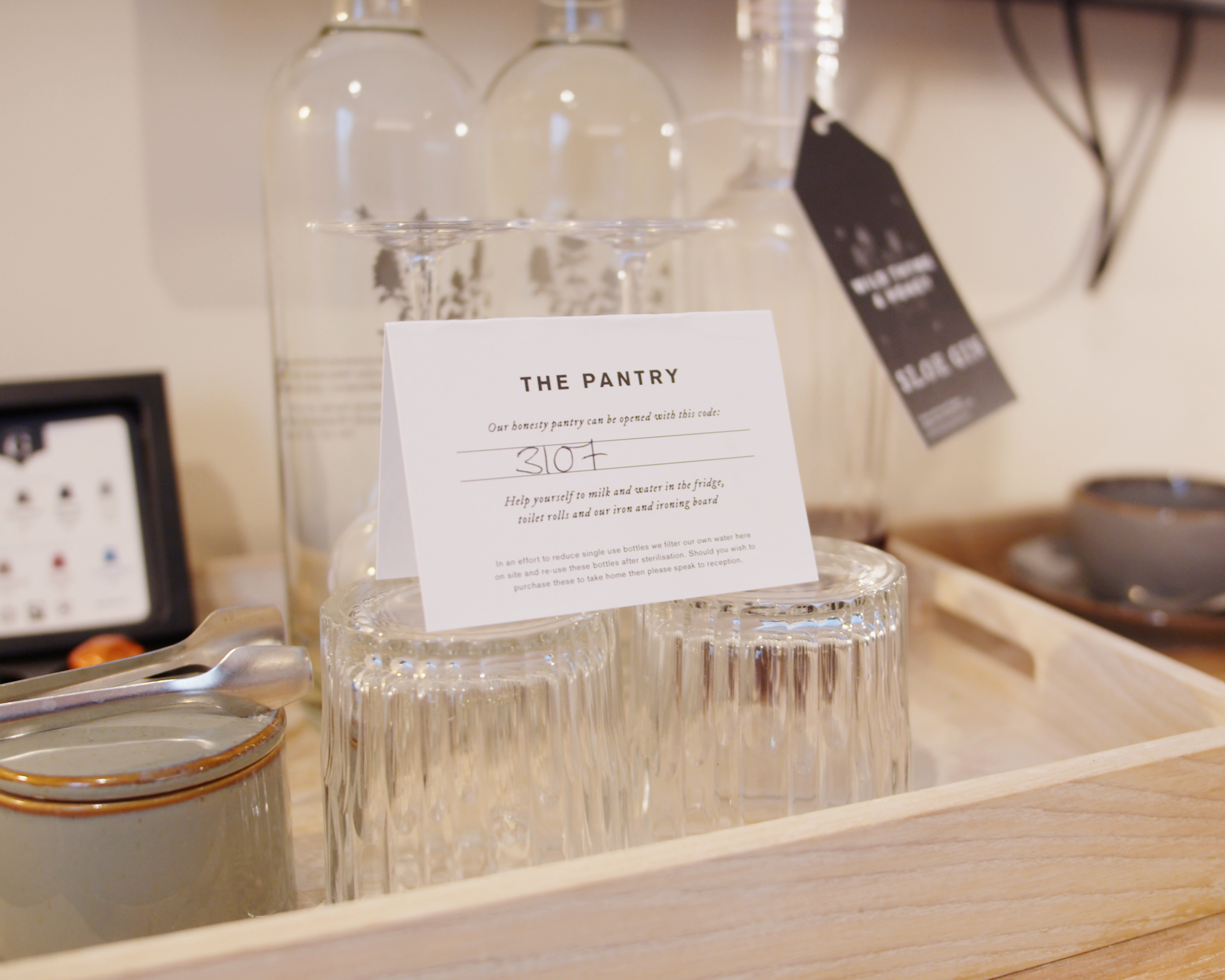 A wooden tray with glass jars, a small ceramic bowl, a tag, and a card labeled The Pantry featuring menu details. - Wild Thyme & Honey Hotel in the Cotswolds