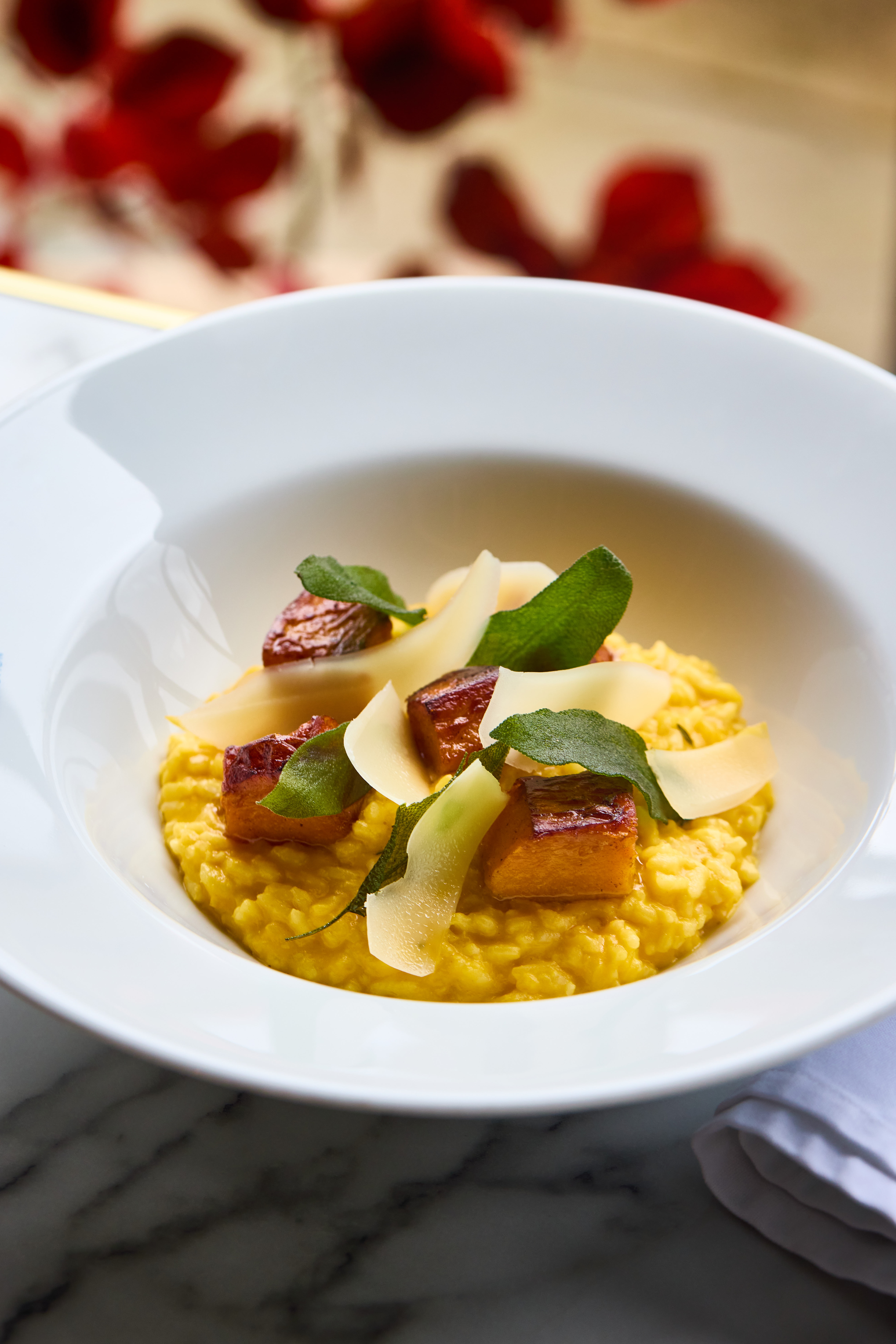 A white bowl with saffron risotto topped with roasted cubes, sage leaves, and cheese shavings on a marble table. - Wild Thyme & Honey Hotel in the Cotswolds
