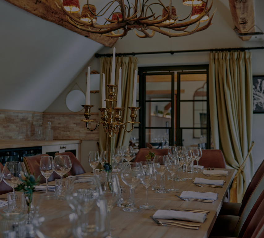Rustic dining room with a wooden table set for a meal, featuring wine glasses, a candelabra, and an antler chandelier. - Wild Thyme & Honey Hotel in the Cotswolds