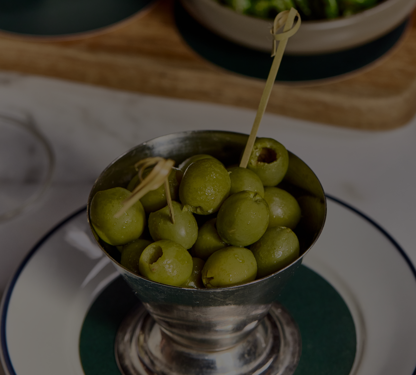 A metal cup filled with green olives, each pierced with a toothpick, on a white and green saucer. - Wild Thyme & Honey Hotel in the Cotswolds