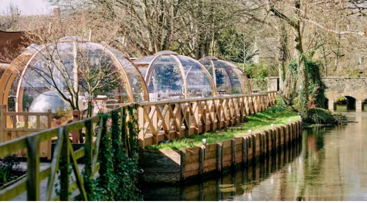 Riverside dining pods with clear domes and wooden frames at The Crown at Ampney Brook, set along a peaceful stream surrounded by trees and greenery. - Wild Thyme & Honey Hotel in the Cotswolds
