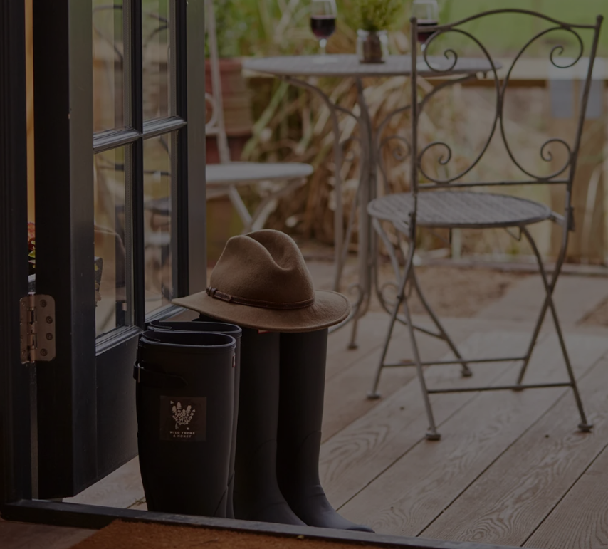 Black rain boots with a brown hat on top by an open door, with an outdoor iron chair and table set in the background. - Wild Thyme & Honey Hotel in the Cotswolds