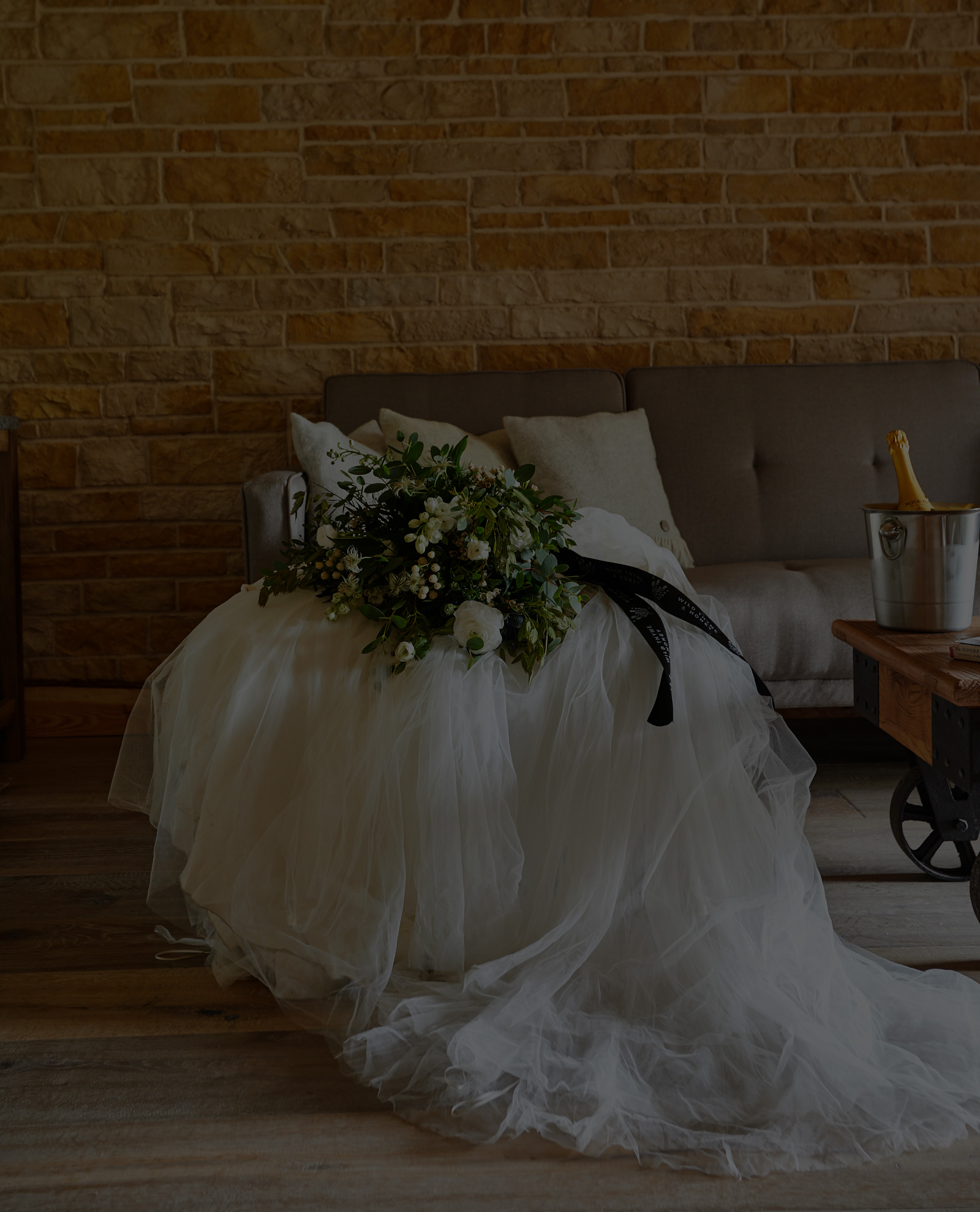 Wedding gown draped over a sofa with a bouquet, next to a wooden table and a champagne bucket against a stone wall. - Wild Thyme & Honey Hotel in the Cotswolds