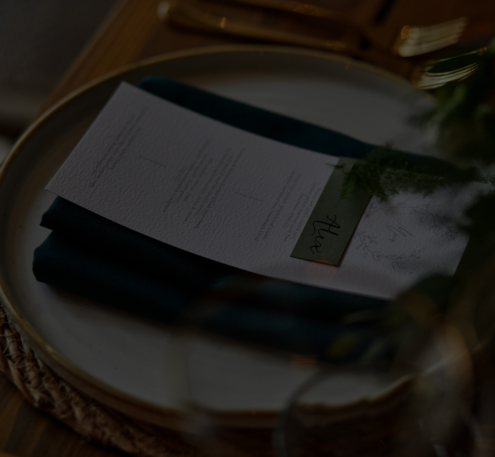 A dinner plate with a folded dark napkin and a menu on top, set on a woven placemat with blurred greenery nearby. - Wild Thyme & Honey Hotel in the Cotswolds