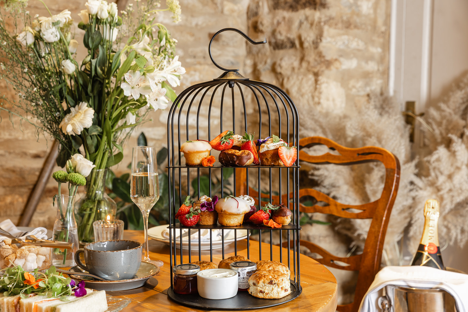 A two-tiered afternoon tea stand with desserts and fruits, surrounded by flowers, tea, and champagne on a wooden table. - Wild Thyme & Honey Hotel in the Cotswolds